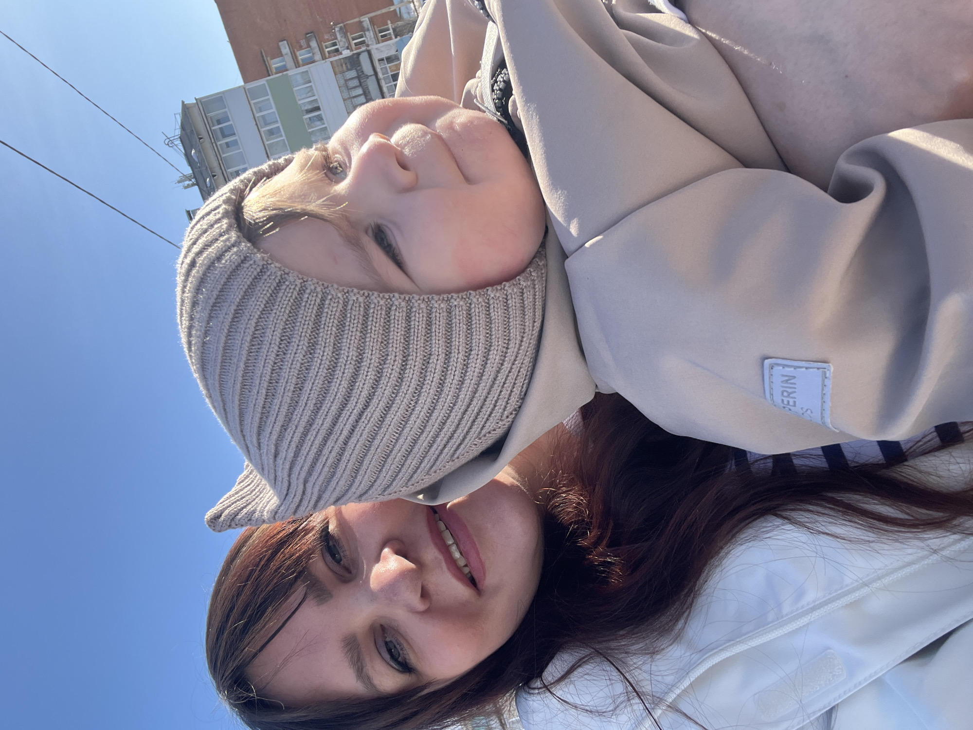 This close-up outdoor image, appearing to be a selfie, shows an adult woman holding a child in the city of Noviy, Russia. The woman, positioned on the left, has long dark brown hair and smiles subtly towards the camera. She wears a light-colored jacket, a white velcro strip visible on its left side. The child, a toddler or infant, is held on the right side of the frame, looking towards the upper right. The child wears a beige ribbed knit hat with ear-like protrusions and a matching beige outer garment featuring a small rectangular label partially legible as "PERIN" and "PS." The background reveals a clear blue sky with two dark utility lines traversing horizontally. Multi-story residential buildings are visible beneath the sky, including one light-colored building with a light green section and another reddish-brown building.