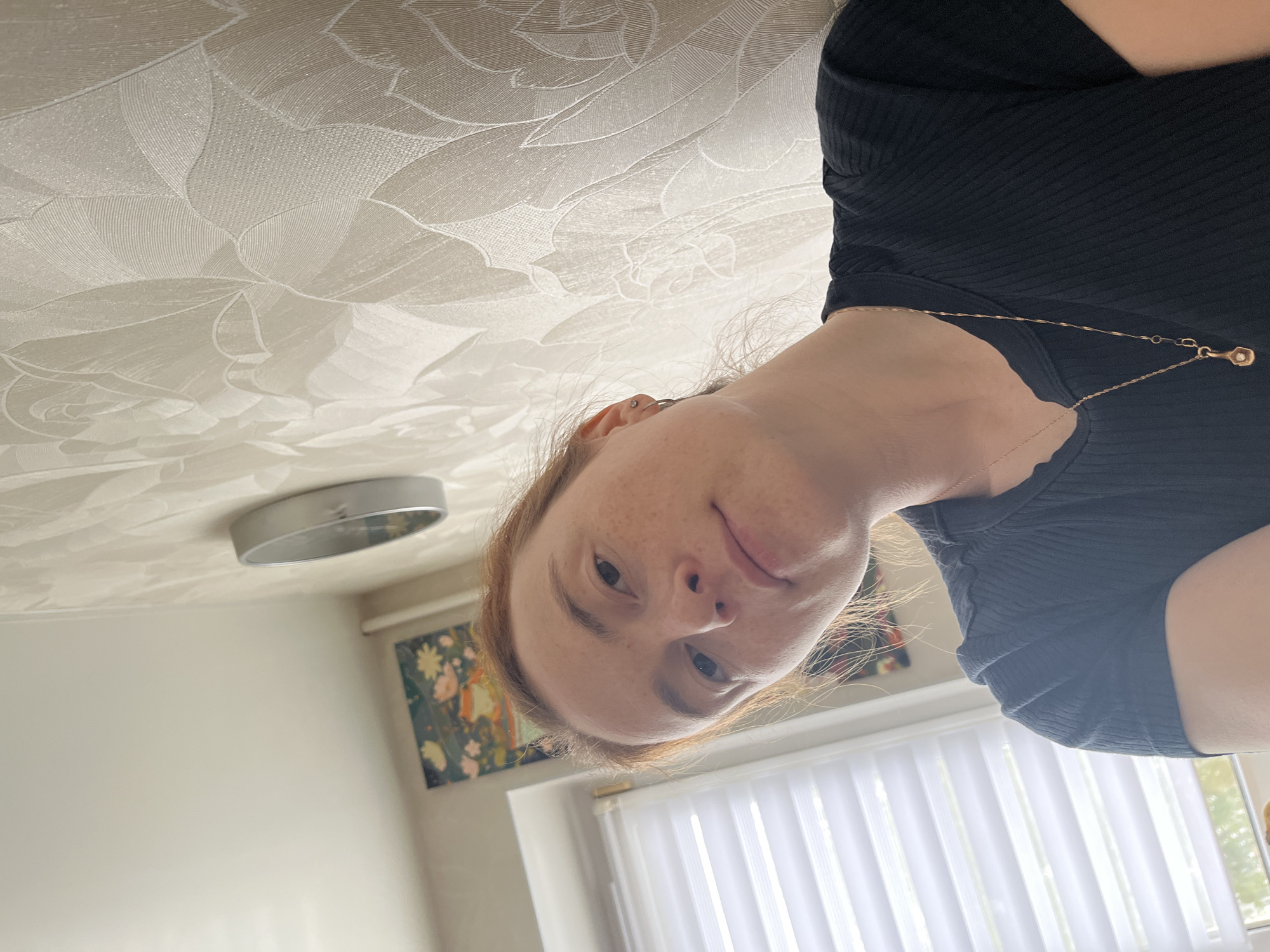 The image features a person, likely female, with fair skin, freckles, and light brown/reddish hair, looking directly into the camera. They appear to be lying down or reclining, with the camera positioned above them. The person is wearing a black ribbed short-sleeved top and a delicate gold-toned necklace with a small, round pendant. A subtle ear piercing is visible on their left ear.
The predominant background element is a white or off-white ceiling, adorned with an embossed or textured pattern of large, stylized floral motifs, resembling roses. A circular, silver-colored light fixture is partially visible on the ceiling towards the upper left of the frame. Below the person's head, in the mid-ground, a section of a white wall is visible. On this wall, a framed piece of art depicting colorful, abstract floral or natural patterns can be seen. Further down, white vertical window blinds are visible, suggesting a window letting in natural daylight.
The scene takes place indoors, likely within a residential setting in Chaikovsky, Russia. The person is posing for the photo, displaying a neutral and relaxed expression. Natural light illuminates the room, indicating it is daytime. There is no visible text in the image.