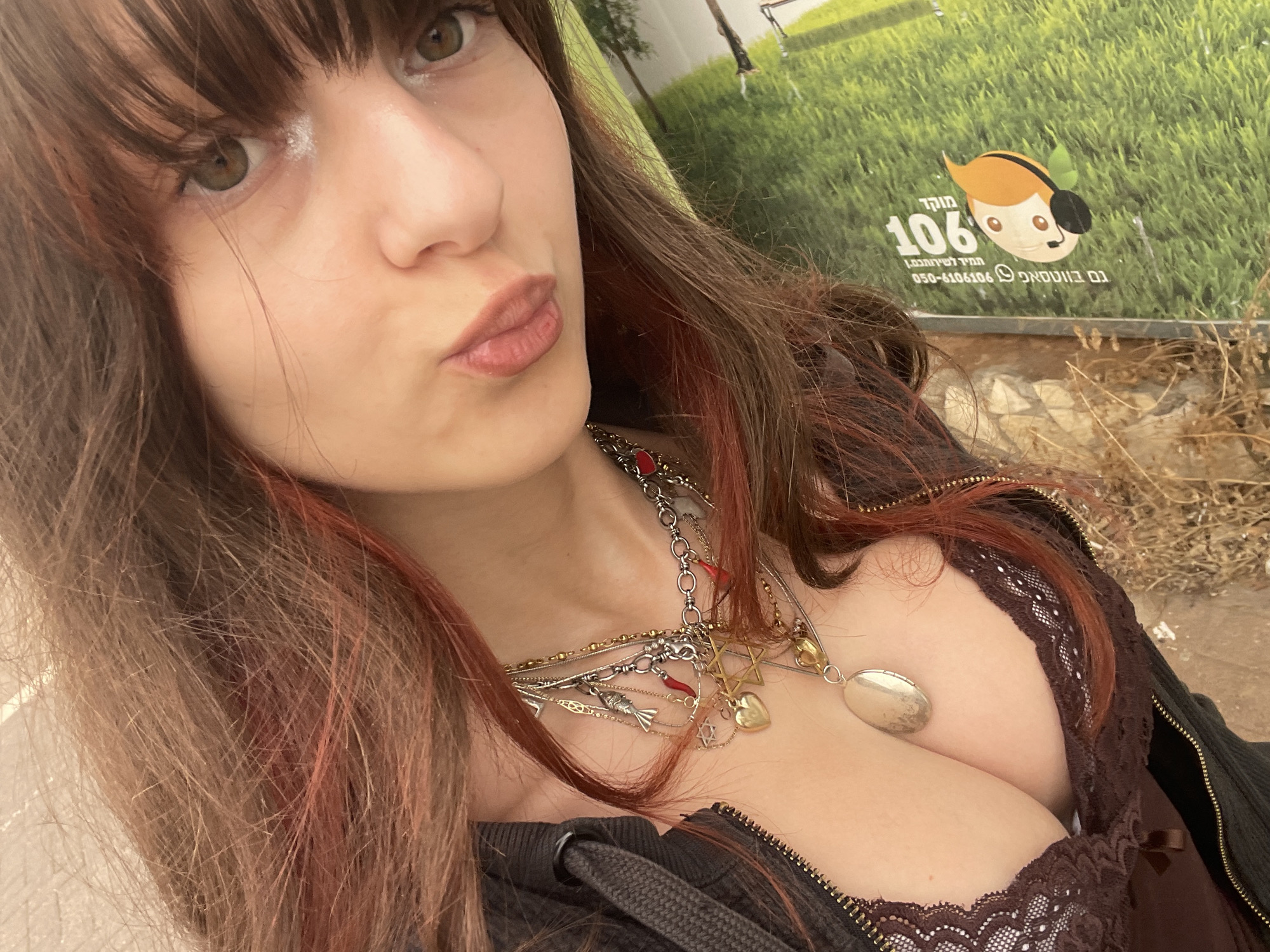 A woman with shoulder-length brown hair with red highlights and bangs is posing outdoors. She is wearing a brown lace top under a dark zip-up jacket, and several necklaces are visible, including one with a Star of David pendant. Her lips are pursed in a kiss. In the background is a sign in Hebrew, featuring a cartoon character and the number "106". Based on the sign, the location is likely in Petah Tikva, Israel. The environment appears to be a public area with some greenery and possibly dried vegetation.