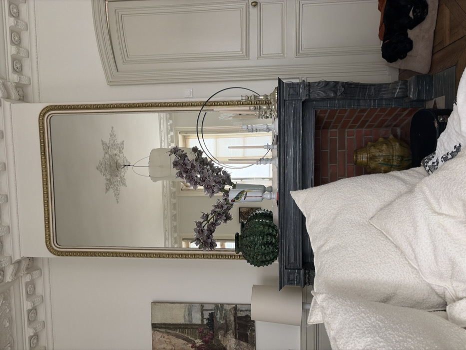 The image depicts an interior scene within a classically designed apartment, characteristic of Haussmannian architecture found in Paris, France. The central feature is a dark-colored, ornate fireplace mantelpiece, likely slate or painted wood, topped by a tall, rectangular mirror with a gold and white beaded frame.
On the mantelpiece, from left to right, are a dark green, textured ceramic vase, a white vase with a colorful bird illustration containing purple orchid stems, several white and silver candelabras holding unlit candles, and a black circular metal decorative object. The fireplace hearth contains red brick and a large, olive-green ceramic pot.
To the left of the fireplace, a framed painting depicting a landscape or still life with muted tones is partially visible, alongside a cream-colored lamp shade. To the right, a tall, white paneled door with a brass doorknob features decorative molding. On the floor to the right of the fireplace, a black dog is resting on a light brown pet bed, partially covered by an orange fabric.
In the foreground, a white, textured sofa or armchair is visible, adorned with several large, fluffy off-white pillows and a patterned throw blanket. The ceiling above the mirror reflects an ornate white ceiling rose and a simple white drum pendant light fixture, along with a glimpse of a room beyond, featuring a window. The overall environment suggests a well-maintained living space with high ceilings and decorative moldings.
