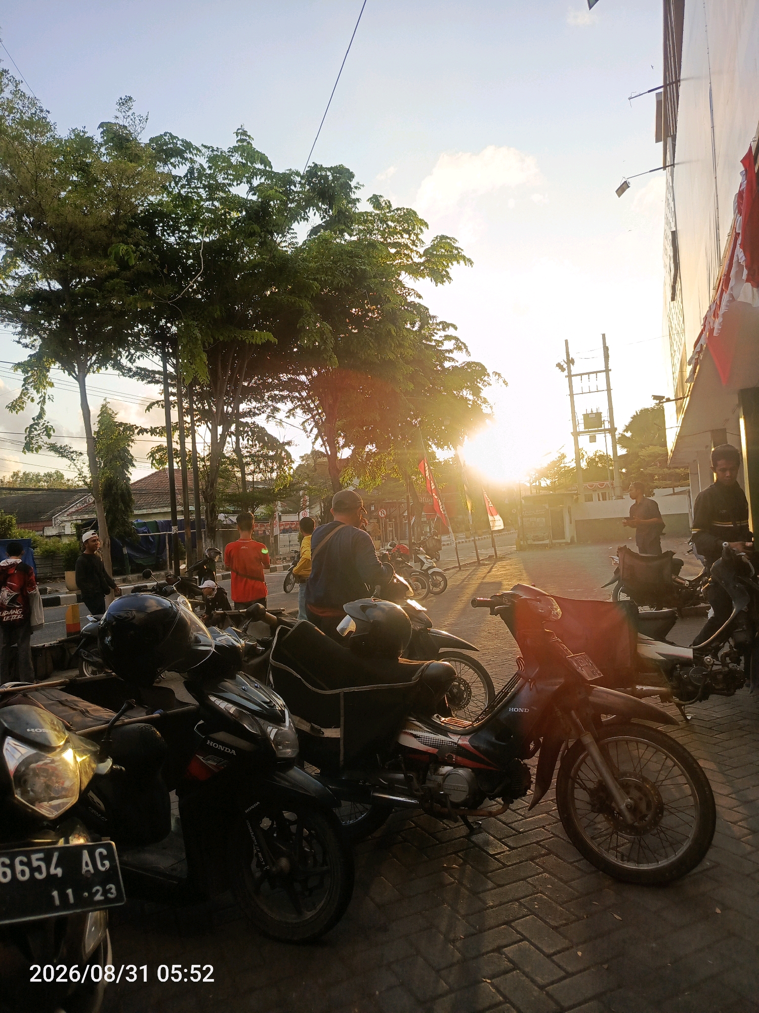 The image depicts a street scene in Pamekasan, Indonesia, likely during the late afternoon or early morning given the golden light and long shadows. In the foreground, several motorcycles are parked on a paved surface. The motorcycles are of various models, with some appearing to be scooters and others more standard models. One motorcycle in the lower left has a license plate that reads "6654 AG 11-23".
A group of people are visible in the mid-ground, some standing around and others appearing to be walking or interacting. They are dressed in casual clothing. One person is wearing a red shirt with a white logo, another a yellow shirt, and a third a dark blue shirt and cap. Behind the motorcycles, there are trees with lush green foliage, some tall utility poles, and buildings, including what appears to be a commercial establishment with red and white flags or banners displayed on its facade. The sun is low in the sky, creating a warm glow and lens flare that partially obscures the background. The overall impression is a snapshot of everyday life in a bustling urban or semi-urban environment in Indonesia.