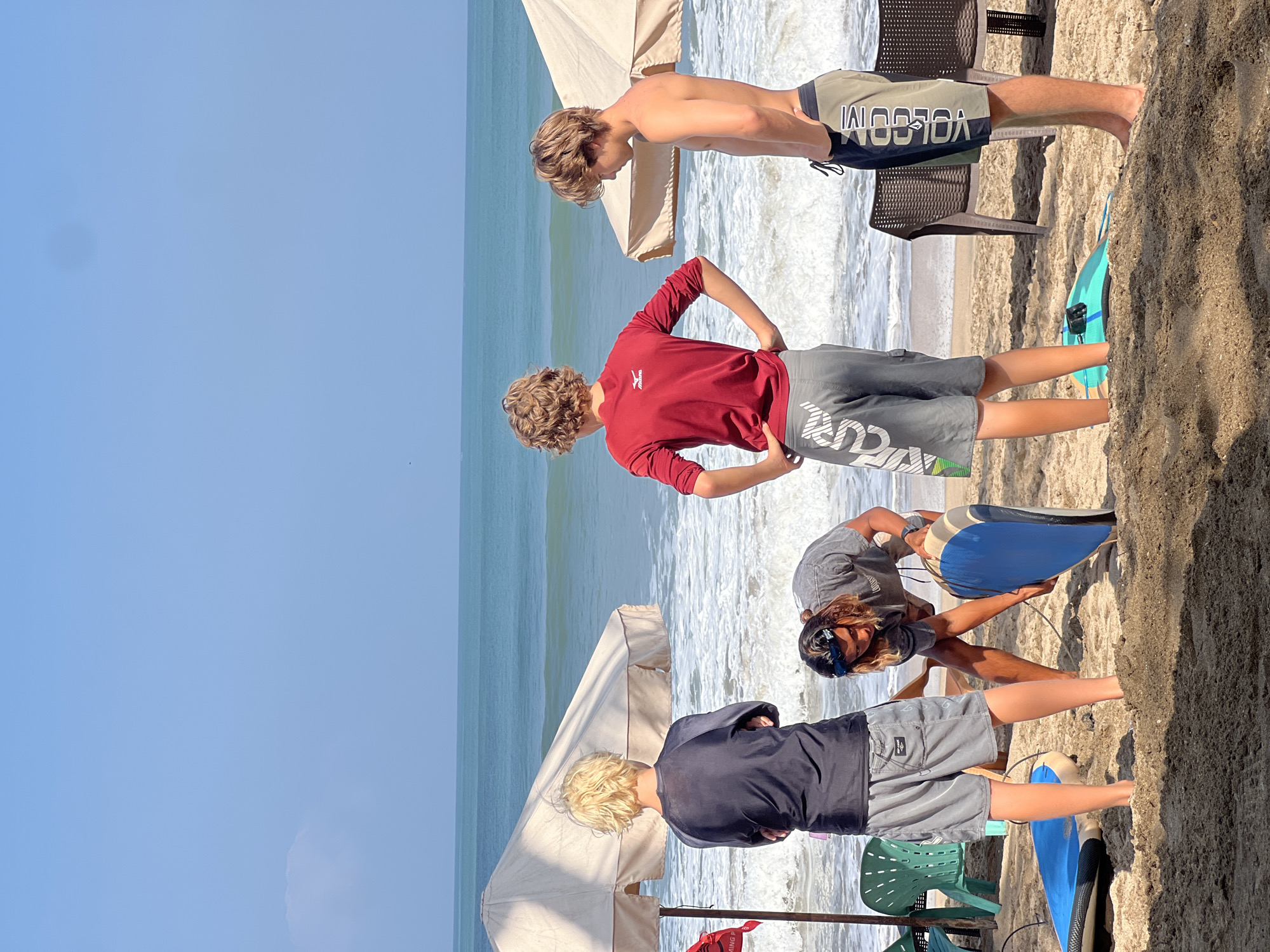 Four young males are on a sandy beach in Kuta, Indonesia, facing the ocean. The sky is clear and blue with a few wispy clouds. The ocean is a bright turquoise with moderate waves. Two large, tan beach umbrellas are set up on the sand, providing shade.
From left to right, the first male is wearing a dark long-sleeved shirt and grey shorts. He is standing with his arms crossed and his back to the camera, looking out at the ocean. Next to him, a second male is bent over, wearing a grey t-shirt and shorts. He is holding a blue bodyboard, appearing to prepare it or pick it up. He has dark, wavy hair and is wearing sunglasses on his forehead.
The third male, in the center, is wearing a red long-sleeved shirt and grey shorts with a white logo. He is standing with his hands on his hips, facing away from the camera and towards the ocean. The fourth male, on the right, is shirtless and wearing khaki and black shorts with a vertical white logo. He is standing with his hands in his pockets, looking down at the sand.
Several surfboards and a small turquoise bodyboard are visible on the sand in the foreground. Beach chairs are scattered behind the umbrellas. The overall atmosphere suggests a group of friends enjoying a day at the beach, possibly for surfing or other water activities.