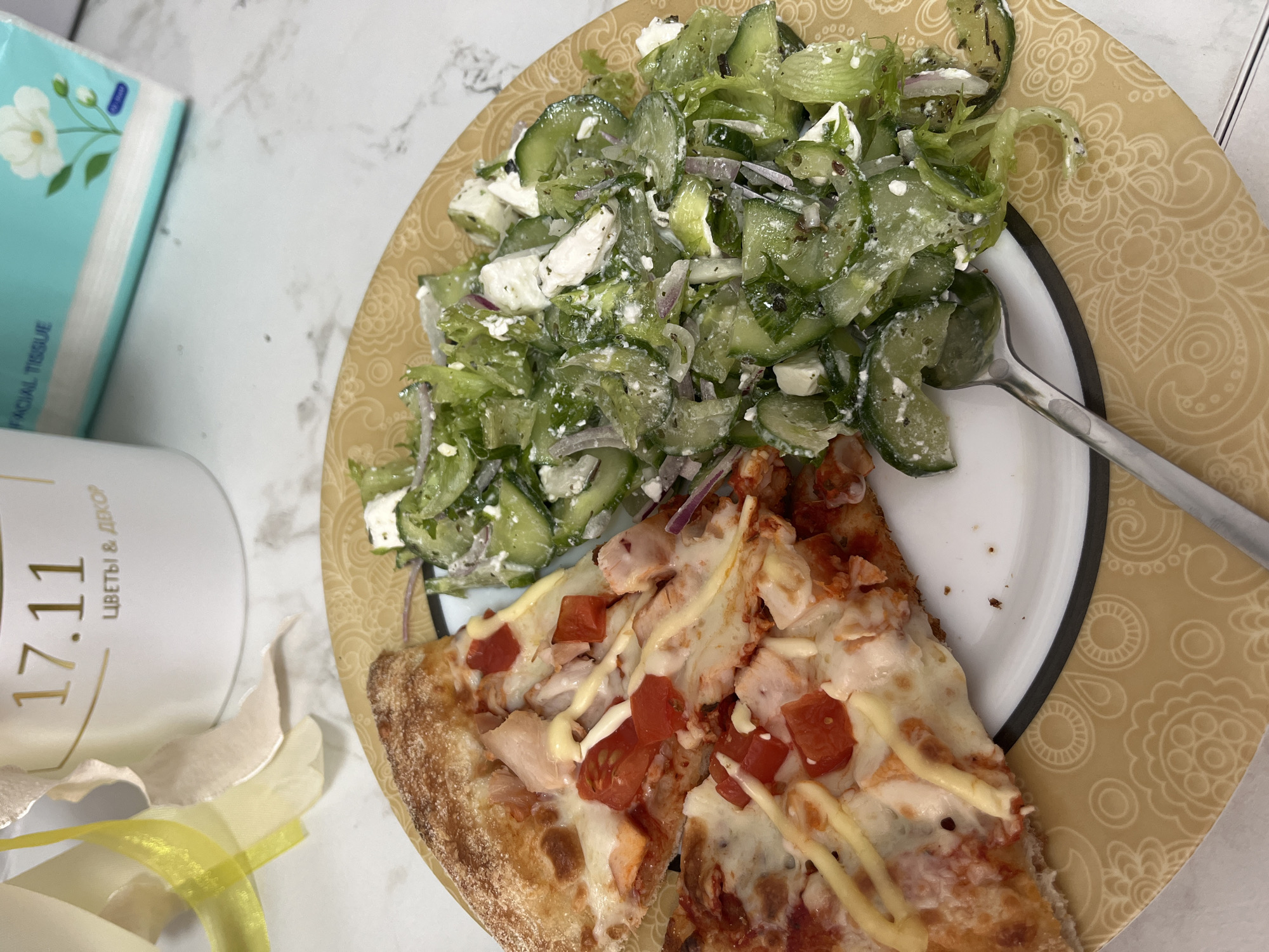 A plate containing a salad and two slices of pizza is the focal point of the image. The salad, positioned at the top of the plate, appears to be composed of cucumbers, lettuce, red onions, and feta cheese. Below the salad, two slices of pizza are arranged, topped with cheese, tomatoes, chicken, and a drizzle of sauce. A metal spoon rests on the plate near the salad. To the left of the plate, there is a box of facial tissues and a container with text in Russian. The container includes the numbers "17.11" and the words "ЦВЕТЫ & ДЕКОР" (which translate to "Flowers & Decor"). The background reveals a light-colored surface, potentially a marble countertop or table. The image could be from Chaikovsky, Russia, based on the Russian text on the container.