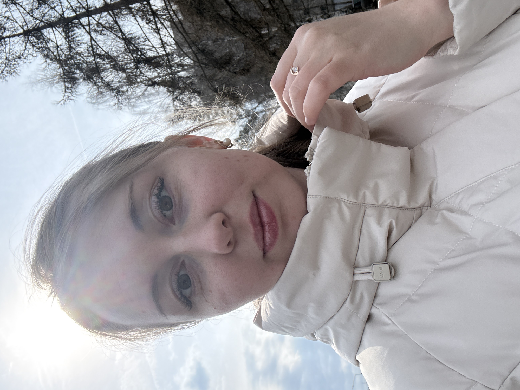 A woman is outdoors in daylight, looking down at the camera. Her face is visible, and she wears a ring on her finger. She is wearing a light-colored, quilted jacket with the hood partially visible. The environment includes bare trees and a sky with some clouds. The location is Unknown, Unknown.