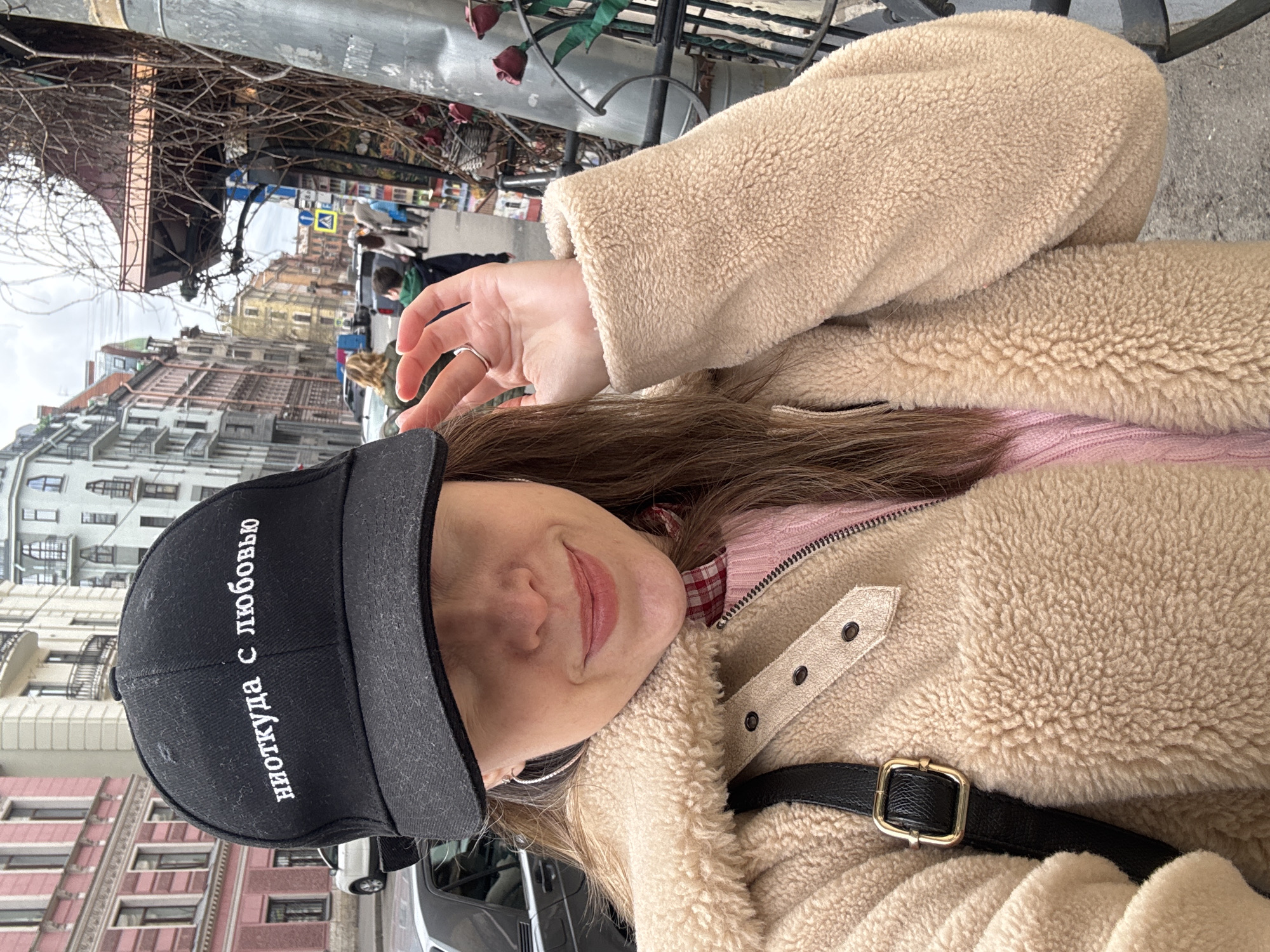 A young woman is captured in a close-up, waist-up shot on an urban street in Saint Petersburg, Russia. The image is taken from a slightly low angle, showing her looking directly at the camera with a soft, closed-mouth smile. She wears a black baseball cap with white Cyrillic text, a beige fleece or teddy bear jacket, and a pink knitted sweater underneath. A black strap from a bag with a gold buckle is visible across her chest. On her left hand, which is slightly raised towards her face, she wears a simple silver ring on her ring finger. Her long brown hair is visible beneath her cap and around her shoulders.
The background reveals a bustling city environment under an overcast sky, suggesting cool weather given her attire. Bare tree branches or overgrown bushes are prominent in the upper left, indicating late autumn, winter, or early spring. Beyond them, a street scene unfolds with multi-story buildings featuring classical architectural details. Distant blurred figures of people and vehicles are visible on the street. A blue and yellow street sign is partially visible in the mid-ground, typical of European road signs. The overall ambiance is casual and urban.
The visible text on her black baseball cap, "ниоткуда с любовью", translates from Russian to "from nowhere, with love."