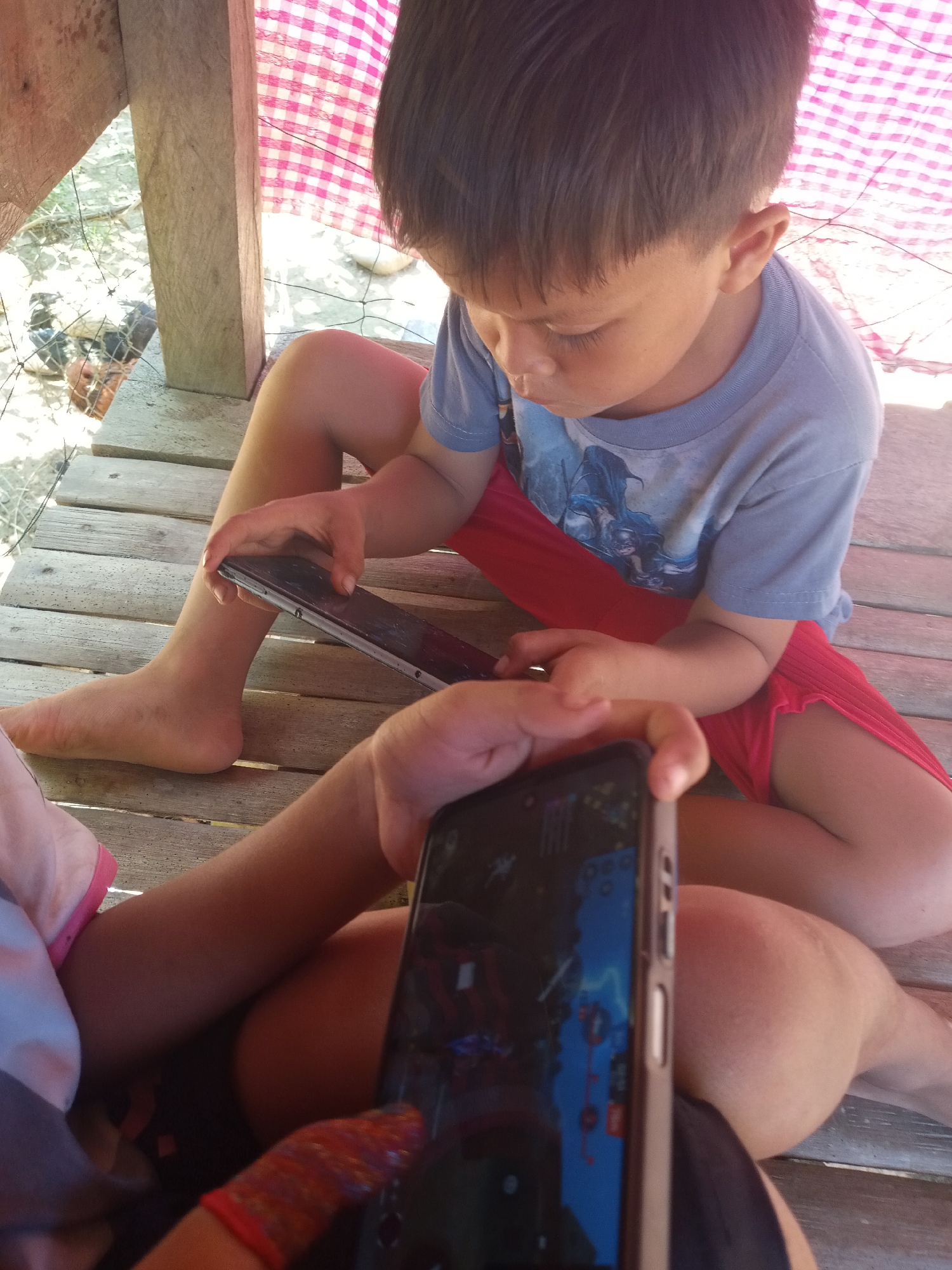 In an outdoor, rustic setting with wooden planks forming a floor and support beams, two children are engrossed with smartphones. The child on the right, a young boy with dark hair, wears a light blue t-shirt with a graphic print and red shorts. He is holding a dark-colored smartphone in his left hand, seemingly focused on the screen. Another child, whose legs and torso are visible in the foreground, also holds a smartphone, displaying a game with colorful graphics. The environment is partially covered by a pink and white checkered fabric overhead. The background appears to be a dirt area with some scattered objects and fencing.