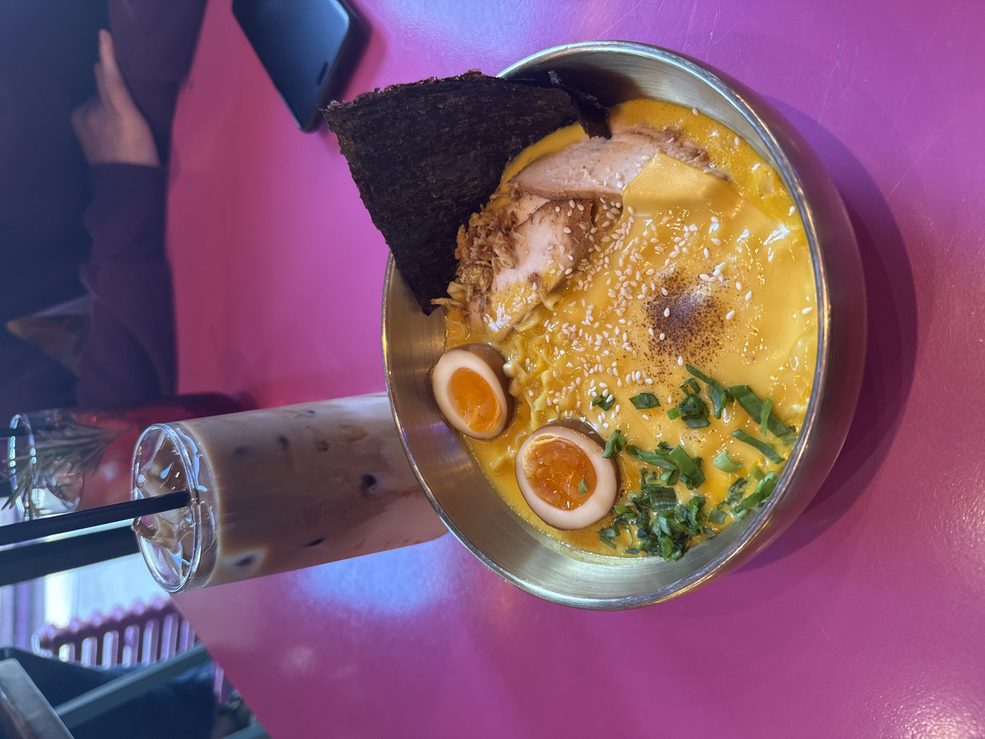 The image shows a bowl of ramen and a glass of iced coffee on a pink table. The ramen contains noodles in a yellow broth, topped with sliced pork, a sheet of nori seaweed, halved soft-boiled eggs, sesame seeds, and chopped green onions. The iced coffee is in a tall glass with a black straw. The image was taken in Moscow, Russia.