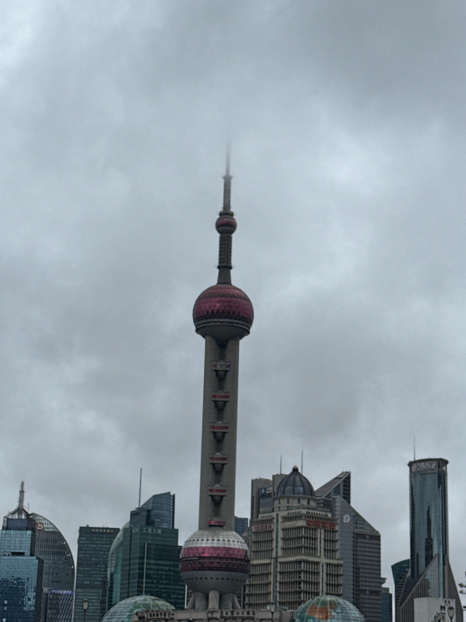 The image depicts a cloudy day in Shanghai, China, focusing on the Pudong skyline. Prominently featured is the Oriental Pearl Radio & TV Tower, a distinctive landmark with its spherical components, partially obscured by mist at its apex. Surrounding the tower are various modern skyscrapers, characterized by their glass facades and geometric designs, typical of the city's business district. The architecture suggests a contemporary urban environment, with no visible individuals or specific actions taking place. The overall atmosphere is subdued due to the overcast sky and diffused light.