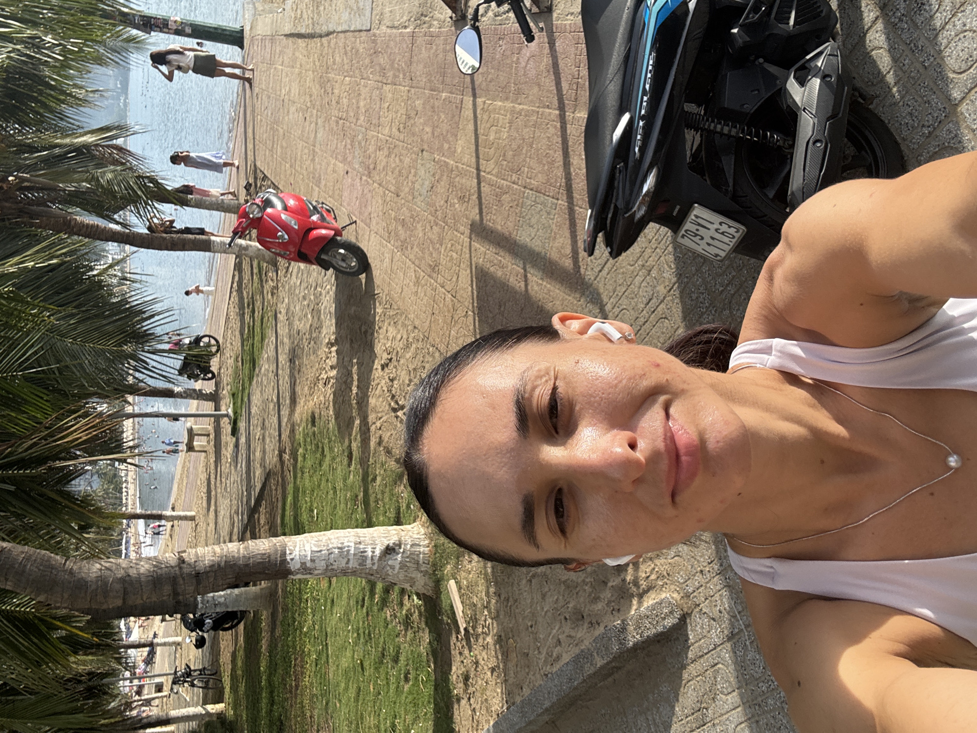 A woman stands on a paved area adjacent to a grassy space and a beach with palm trees in Bac Nha Trang Ward, Vietnam. She wears a white tank top and a necklace and has earbuds in her ears. A black and blue motorcycle and a red scooter are parked nearby. People are visible on the beach. The license plate on the motorcycle reads "79-Y1 411.63." The weather appears sunny and bright.