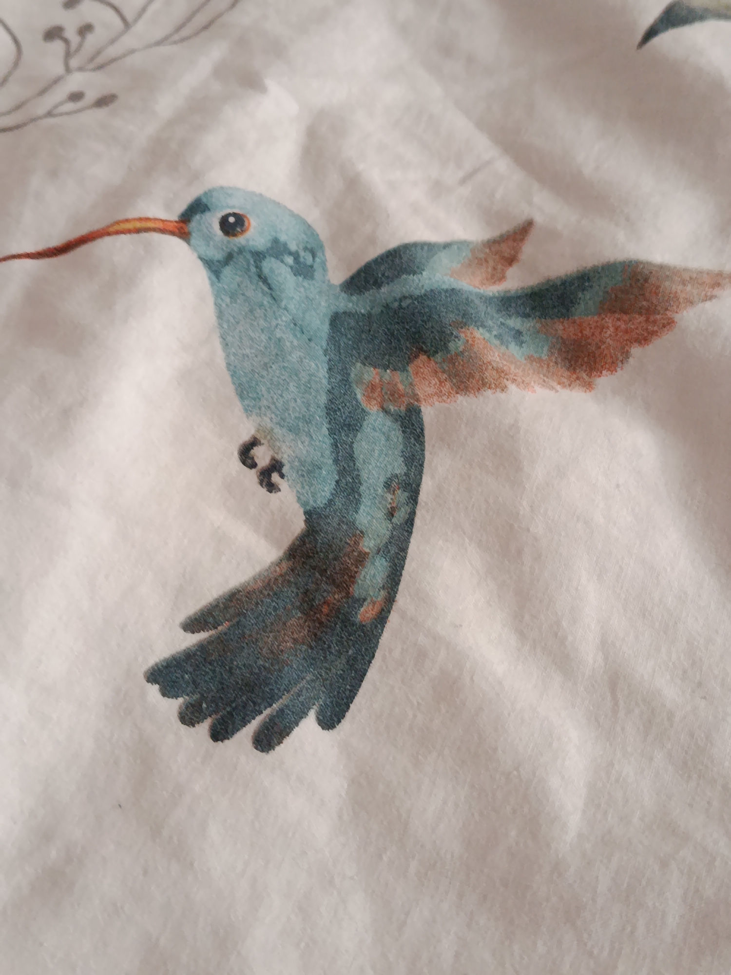 The image shows a close-up of a white fabric with a painted illustration of a hummingbird. The hummingbird is depicted in flight, with its wings spread and its beak extended. It is predominantly blue and teal, with accents of rust and brown on its wings and tail. The fabric has a slightly textured appearance, and the painting style is somewhat painterly, with visible brushstrokes. There are no people or identifiable landmarks visible in the image. The image does not provide any information about the city of Svietlahorsk, Belarus.
