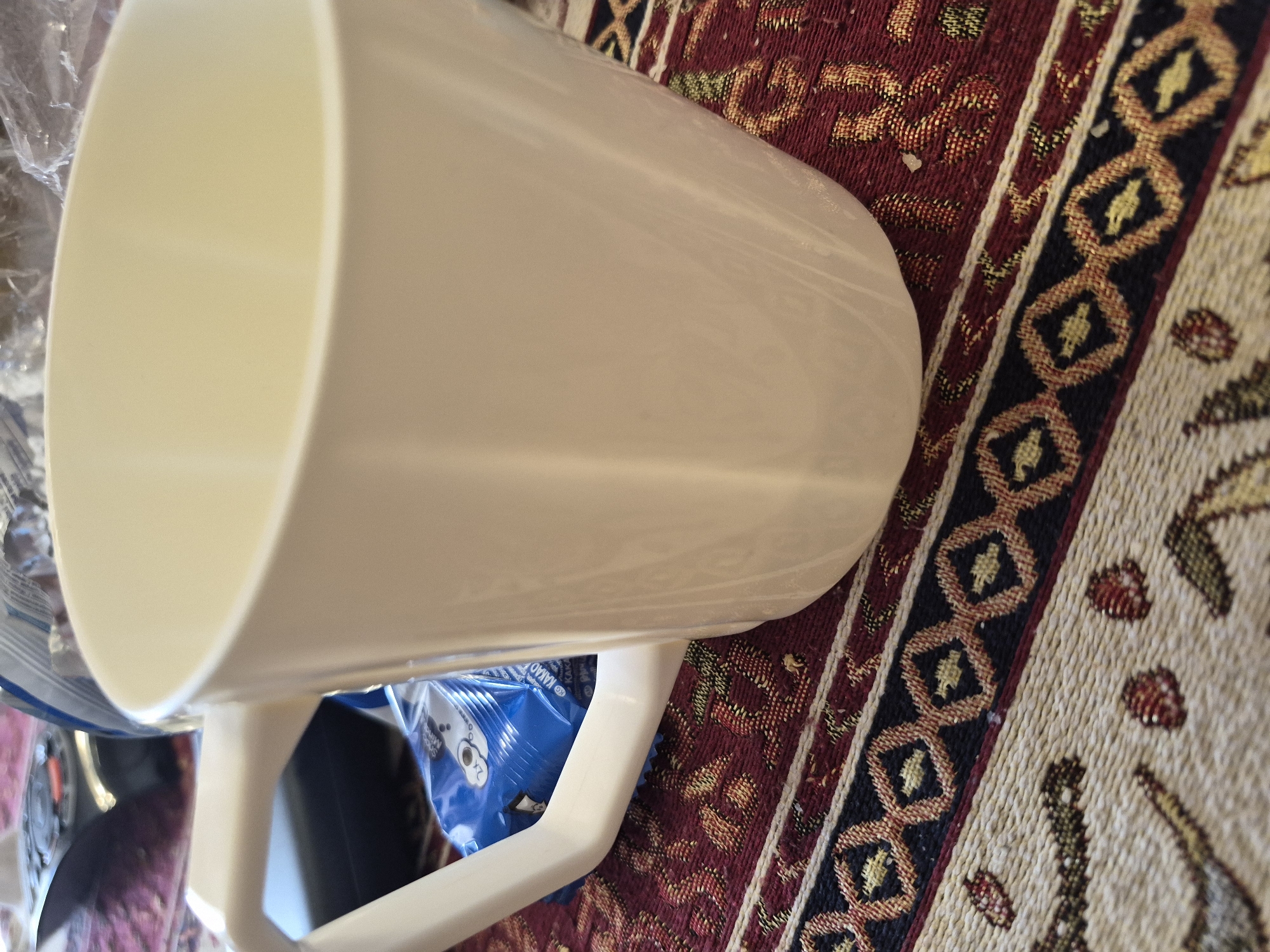 The image displays an off-white or cream-colored mug lying on its side, positioned across the center and left of the frame. The mug, which appears to be made of plastic or ceramic, has its handle visible towards the bottom-left.
The mug rests on a richly patterned rug or textile. The rug features prominent bands of deep red adorned with golden and green accents, and a distinct dark blue stripe showcasing a repeating pattern of interconnected golden diamond shapes. To the right, a lighter beige section with smaller, stylized red and green motifs is visible. The woven texture of the rug is clearly evident.
Partially visible beneath and behind the mug is a crinkled blue plastic package. Fragmented text on this packaging includes "KAKAO" and "Smooth M", along with a "2x" notation. An illustration resembling two circular cookies or cartoon eyes is also present, indicating the package likely contains a cocoa-flavored snack.
The scene is an indoor setting. No human subjects or explicit activity are depicted; the objects are static on the patterned surface. No specific time of day or emotional context can be determined.