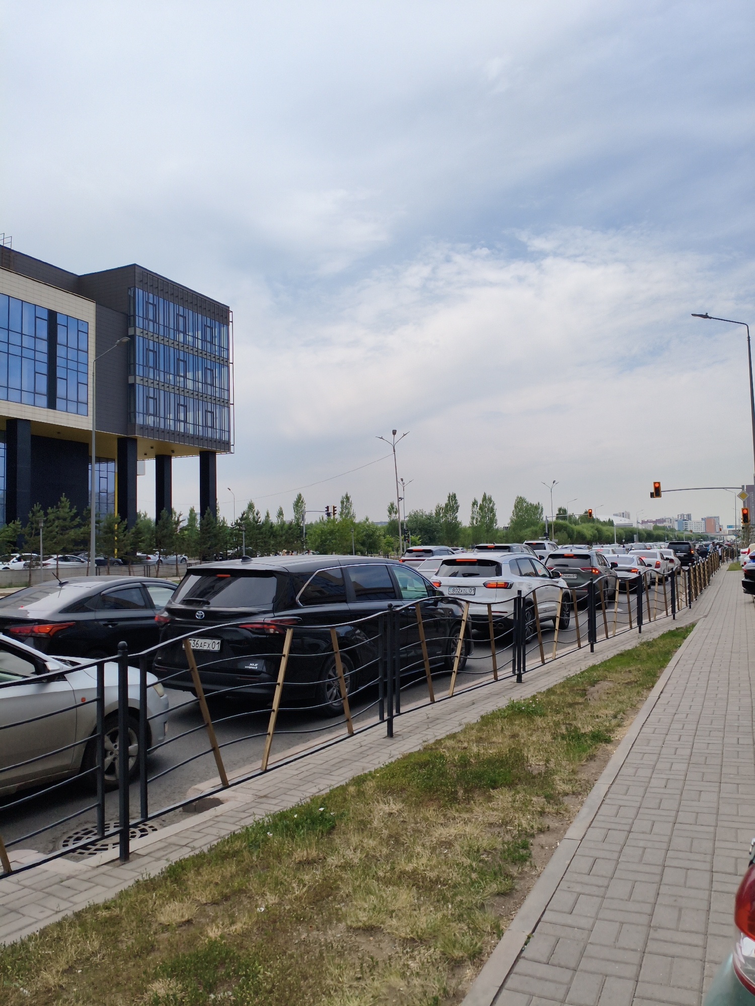 The image depicts a street scene in Astana, Kazakhstan, characterized by heavy traffic congestion. A modern building with a glass facade stands prominently on the left, accompanied by streetlights. Multiple cars are lined up on the road, indicating a traffic jam. A sidewalk made of paving stones runs along the right side of the road, bordered by a grassy area and a black metal fence. The sky is partly cloudy.