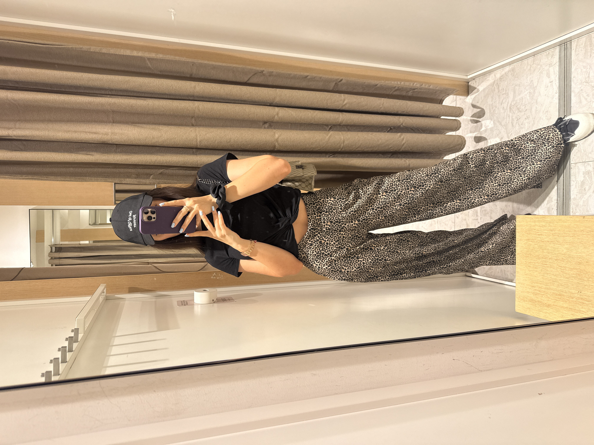 The image depicts a person taking a photo of themselves in a fitting room, likely in a clothing store. The individual is wearing a black t-shirt, leopard-print pants, and a black baseball cap. They are holding a purple smartphone to take the picture. The fitting room has a mirror, clothes racks, and a curtain in the background. The location is not evident from the image; therefore, identifying Plovdiv, Bulgaria, is not possible.