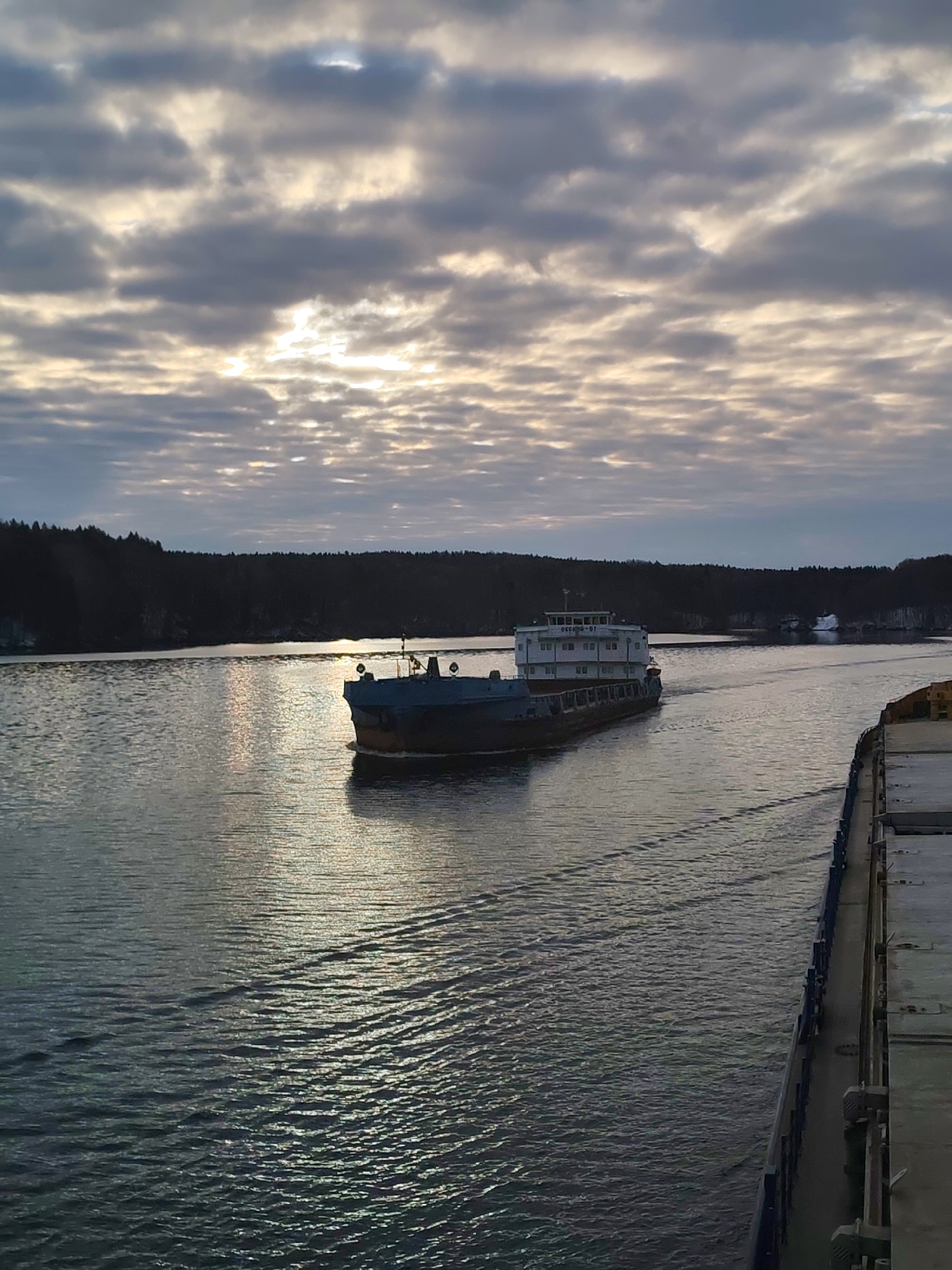 This media file captures a serene scene on a wide waterway, likely a river or canal, under an overcast sky with patches of sunlight breaking through. The time appears to be daytime, possibly morning or late afternoon, given the soft, diffused light.
The primary subject is a large, grey cargo ship or barge, marked with the text "БЕСКРАЙ - 07" (BESKRAY - 07) on its white superstructure, sailing towards the viewer, slightly left of the center. It leaves a distinct wake behind it on the calm water. To the right of the frame, the fore or aft section of another large vessel, from which the photo is presumably taken, is visible, showing its deck and side hull.
The waterway is flanked by dense, dark evergreen forests on both sides, suggesting a cold climate or time of year. The sky is predominantly covered by a layer of altocumulus or stratocumulus clouds, allowing beams of sunlight to illuminate parts of the clouds and reflect brightly on the water surface, creating shimmering patterns. The overall atmosphere is tranquil and slightly muted in color. The location context of Ozerki, Russia, suggests this is an inland waterway, possibly part of a river system or canal network in the region.
