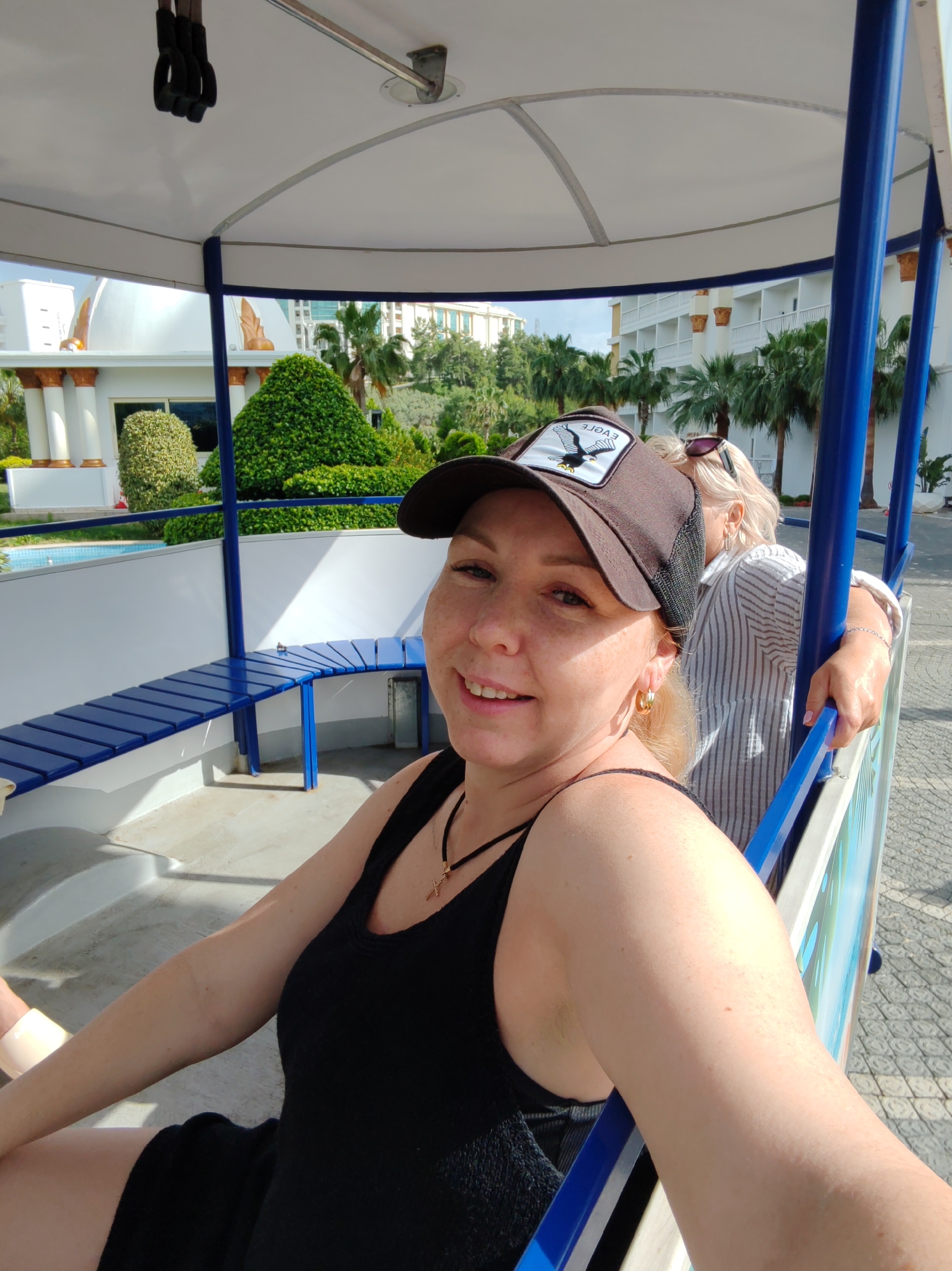 A woman is taking a selfie while riding in a vehicle with blue benches and support poles. The vehicle has a white roof. The woman wears a black tank top, a brown cap with an eagle design, and gold hoop earrings. She has a necklace with a cross pendant. Another person with blonde hair is partially visible behind her, wearing sunglasses and a striped shirt. The vehicle is moving past a building with white walls, columns, and decorative gold features. There are manicured green bushes and palm trees in the background. The location is Alanya, Turkey, as indicated by the architectural style.