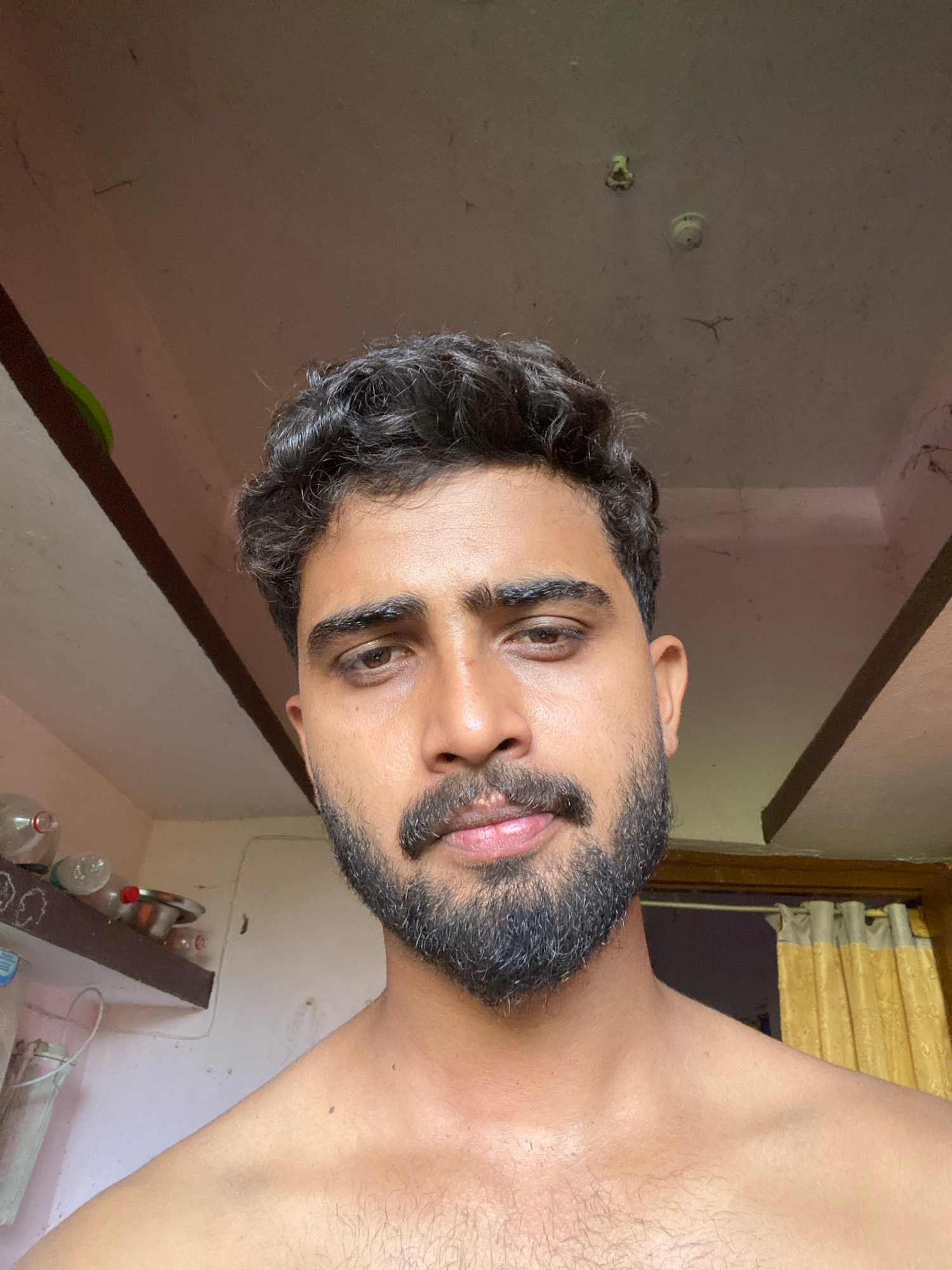 A close-up, waist-up photograph taken from a low angle shows a young man in Bahadurpur, India. He has dark, curly hair, a thick beard, and is looking directly at the camera with a slight smile. His chest and shoulders are bare. The background shows a room with a textured ceiling, wooden beams, shelves with bottles and bowls, and yellow curtains. The lighting is soft and natural.