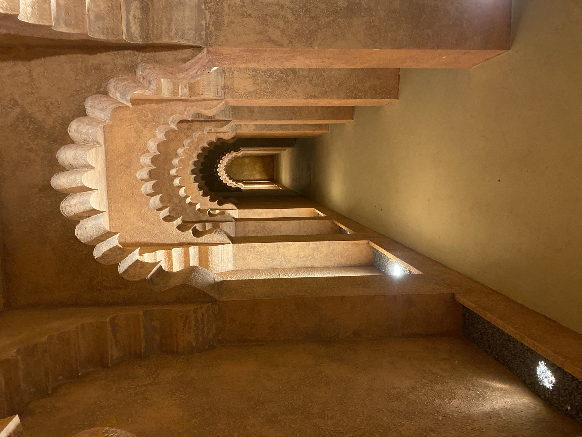 This is an interior shot of a staircase in Mũi Né, Vietnam. The structure is primarily constructed from a sandy-brown material, possibly concrete or stone. The staircase has a decorative, scalloped edge along the outer side. The steps descend into a narrow, tunnel-like passage illuminated by small recessed lights along the right wall. The walls are a muted yellow color. The scene appears to be enclosed and artificial lighting is used. The perspective is looking down the staircase.