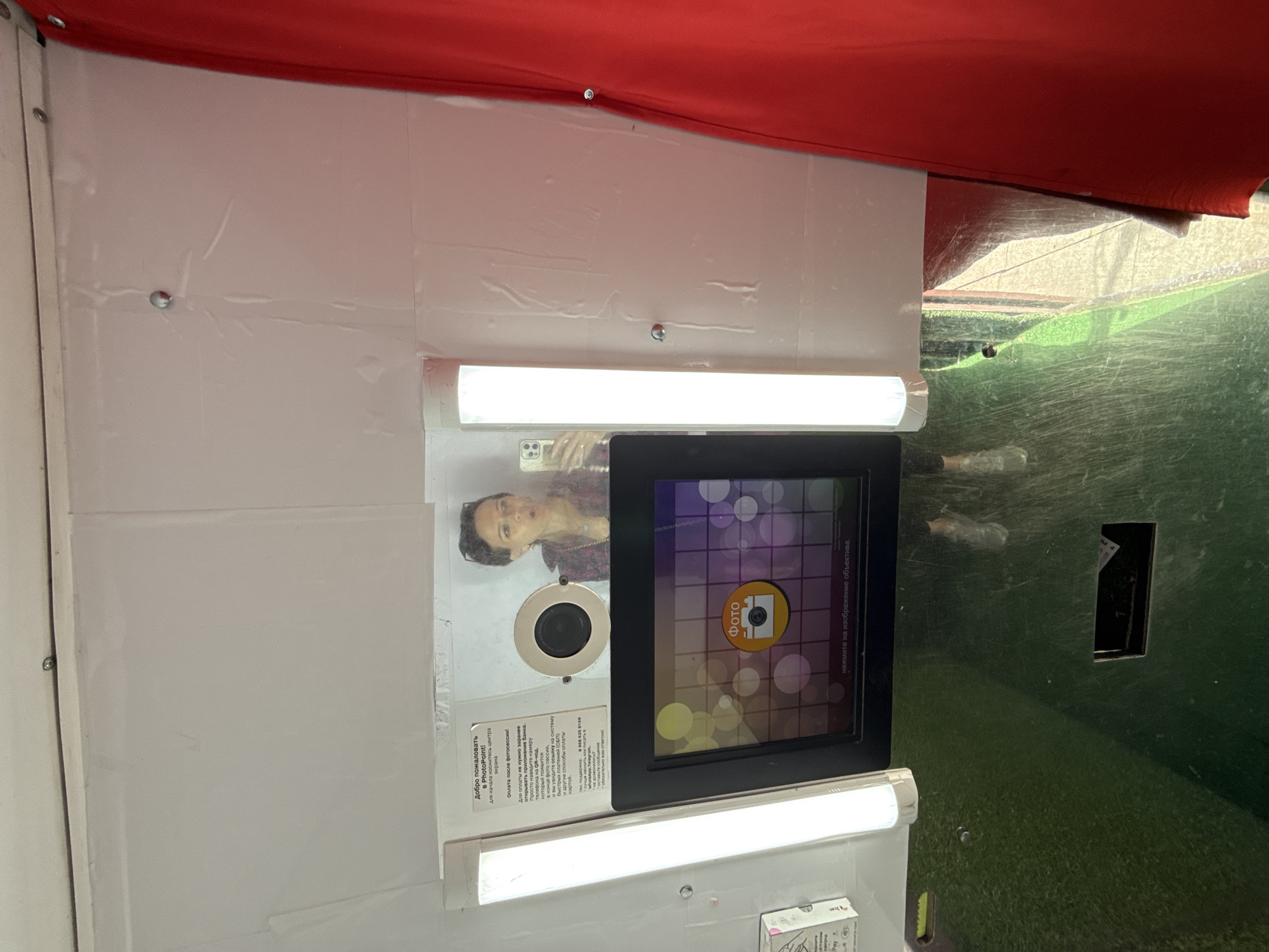 This media file shows the interior of a photo booth. A woman is visible in a reflection on the screen of the booth. The screen displays a "Photo" icon with a camera symbol and animated circles. There are two bright fluorescent lights illuminating the space. A red fabric canopy hangs overhead. To the right of the photo booth, a green, textured surface is visible, suggesting it might be outdoors or in a space with a green wall. A small black rectangular opening is present on this green surface. Text in Russian is visible on a sticker attached to the photo booth, which translates to "Welcome to PhotoPoint! To start, touch the center of the screen. Payment after the photoshoot! To pay after the photoshoot, you don't need to open the bank app in advance. Just point your phone camera at the QR code that will appear at the end of the photoshoot, and you will see a link to the faster payments system (SBP) and other card payment methods. Technical support: +7 968 625 8168. It's better to call or write on WhatsApp, Telegram. Leave a message - we will definitely reply!" This suggests the photo booth is located in Moscow, Russia. The lighting indicates it is likely daytime.