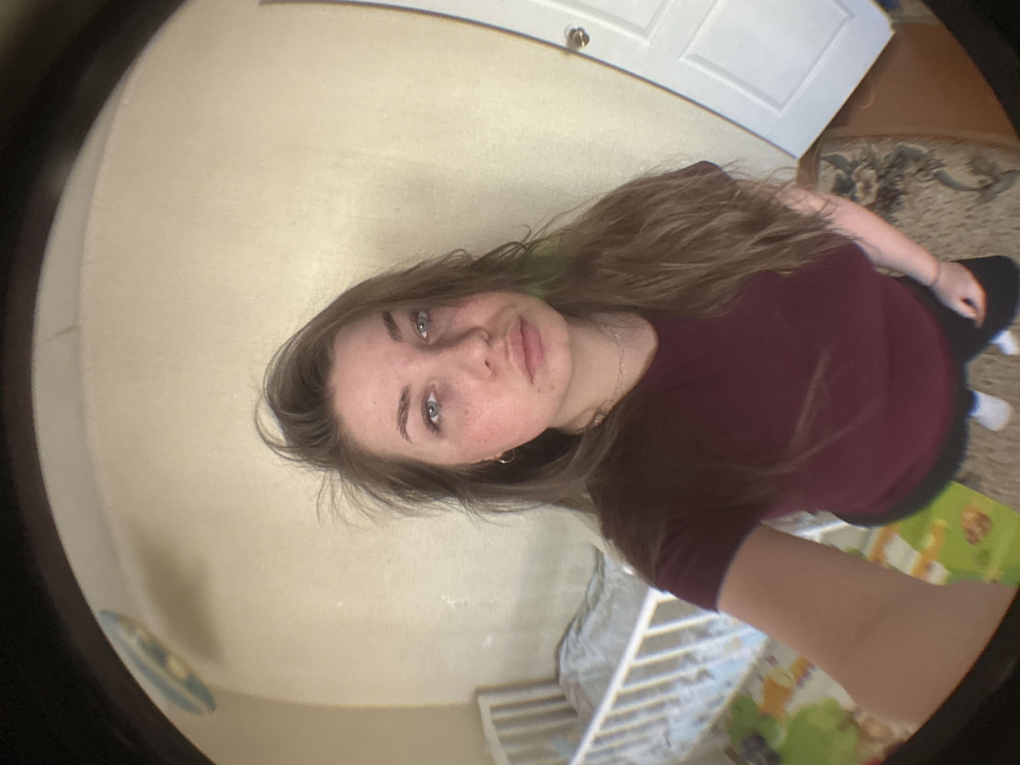 A fisheye lens captures a selfie-style image of a fair-skinned young woman with long, light brown hair. The woman has blue eyes and freckles. She is wearing a dark reddish-purple shirt and a silver necklace.
The background includes a cream-colored wall and a white door with a silver doorknob. Parts of a patterned rug and a white baby crib are visible. The room appears to be inside a home, possibly located in the city of Chaikovsky, Russia. No specific details confirm this, but given the request for the city, it is assumed.