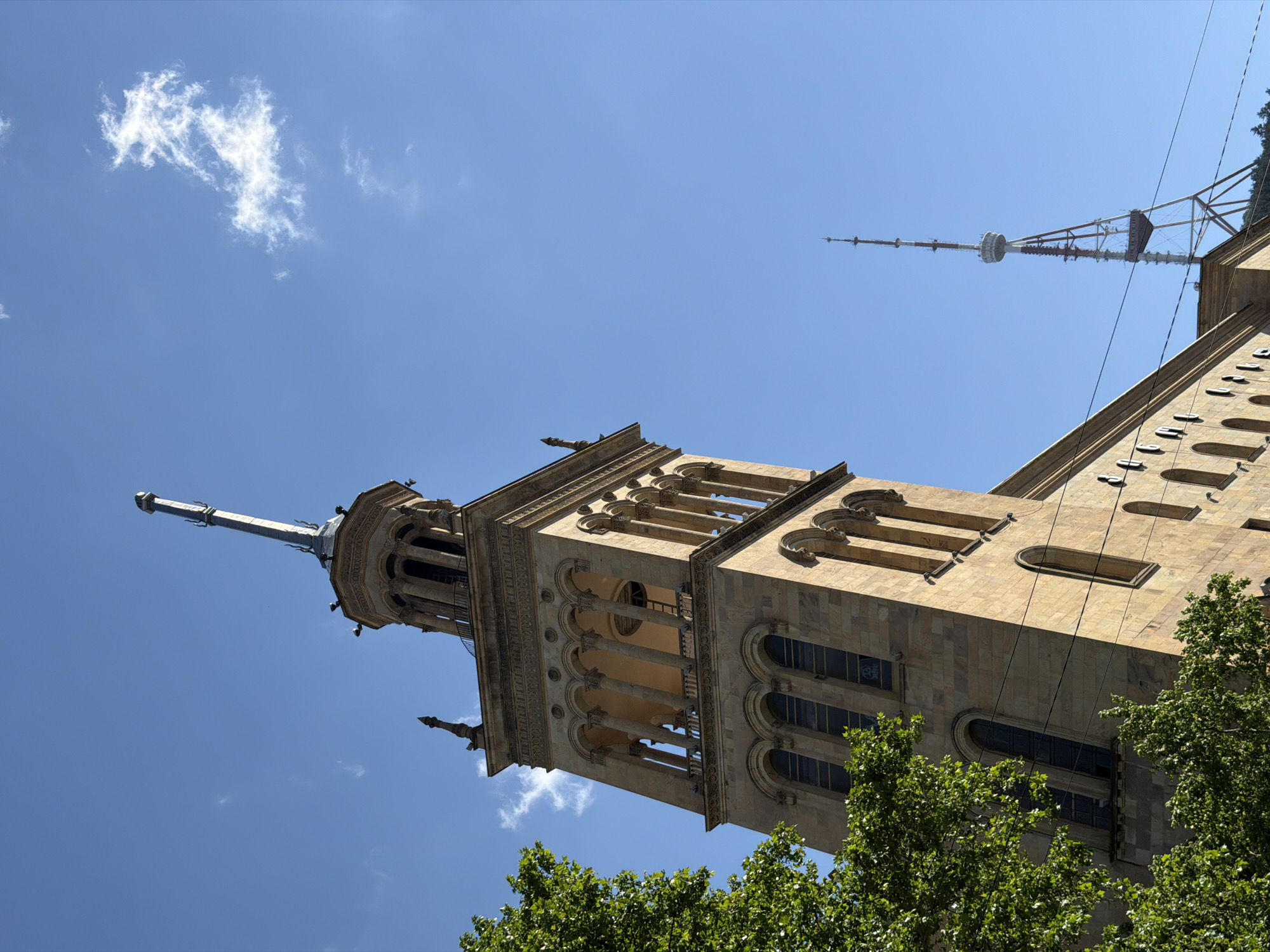 The image shows a low-angle view of a building in Tbilisi, Georgia, specifically focusing on the bell tower of the Tbilisi City Assembly. The beige stone building features arched windows, pillars, and decorative elements. A metal spire rises from the top of the tower. In the background is the Tbilisi TV Broadcasting Tower. The sky is blue with a few scattered clouds. The top of green tree foliage is visible at the bottom of the frame.