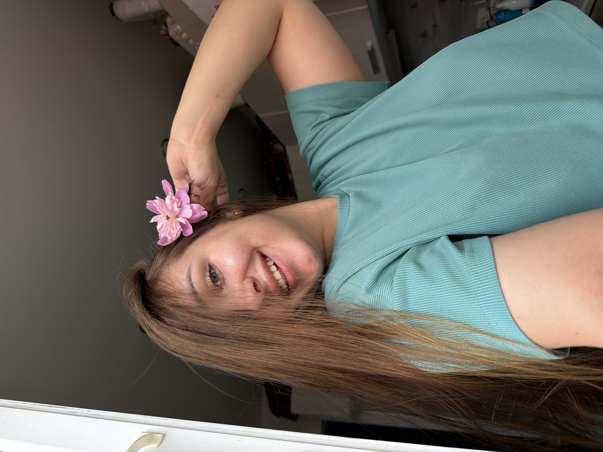 A young woman with long brown hair and a pink flower tucked behind her ear smiles directly at the camera. She is wearing a teal, ribbed t-shirt. Her left hand is raised behind her head, supporting the flower. The background is blurred, showing hints of a room with shelves and bottles. The scene is likely indoors, possibly in a domestic setting. No specific location in Chaikovsky, Russia, can be identified from the visual information provided.