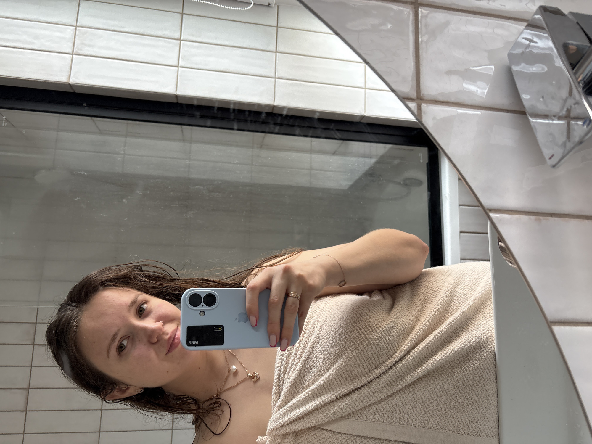 A young woman is taking a selfie in a bathroom. She is holding a light blue iPhone with two camera lenses. The woman is partially covered in a light beige towel. The bathroom has white rectangular tiles on the wall. There is a shower with a black frame visible through the mirror. A chrome faucet is partially visible in the upper right corner of the mirror. The image does not show enough information to establish the location in Izhevsk, Russia.