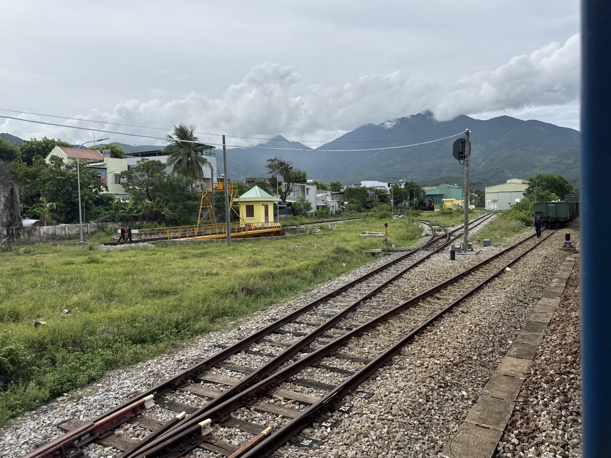 The image captures a rural train yard scene with a backdrop of cloudy, mountainous terrain. Multiple sets of railway tracks dominate the foreground and midground, laid on a bed of gravel. To the left, a grassy area separates the tracks from a collection of buildings, including houses and a yellow structure with a yellow ladder. Further back, industrial-looking buildings and a green train car are visible, along with a single person walking near the train car. The sky is overcast with substantial cloud formations, and the distant mountains are partially obscured by mist. The scene appears to be in a developing area with a mix of residential, industrial, and transportation infrastructure.
