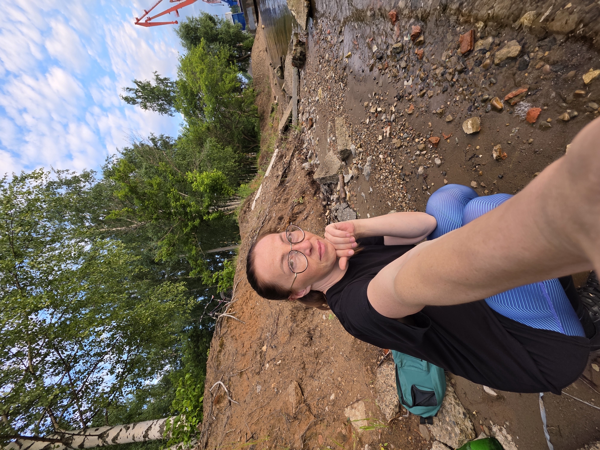 The media file is a selfie taken by a person with long brown hair and glasses. They are wearing a black t-shirt and blue striped leggings. The person is lying on a rocky, muddy shore with a body of water to their right. In the background, there are trees and a blue sky with white clouds. A red crane is visible in the upper left corner, suggesting an industrial area or a port. A green backpack is on the ground next to the person. The image appears to be taken during the daytime. The person has a relaxed expression, resting their chin on their hand. The location context indicates Chaikovsky, Russia, which is a city in Perm Krai, situated on the Kama River.