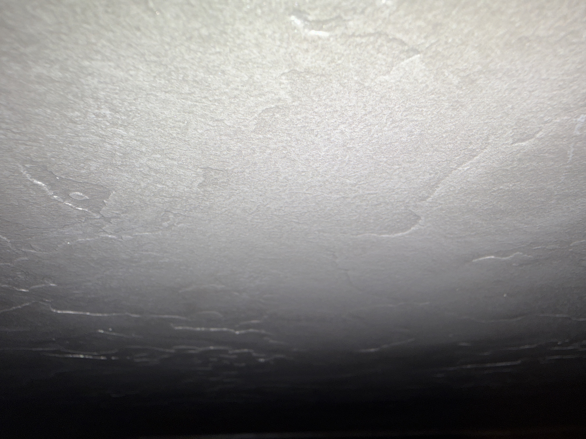 The image shows a close-up, side-view of a textured wall with a gradient of lighting. The left side of the image is dark, fading into a lighter gray and then to a bright, almost white textured surface on the right. The texture appears to be consistent throughout the visible area and resembles a coarse stucco or Venetian plaster. There are subtle, irregular patterns within the texture, suggesting the application technique of the material. No people, objects, or environmental details of a city are visible.
