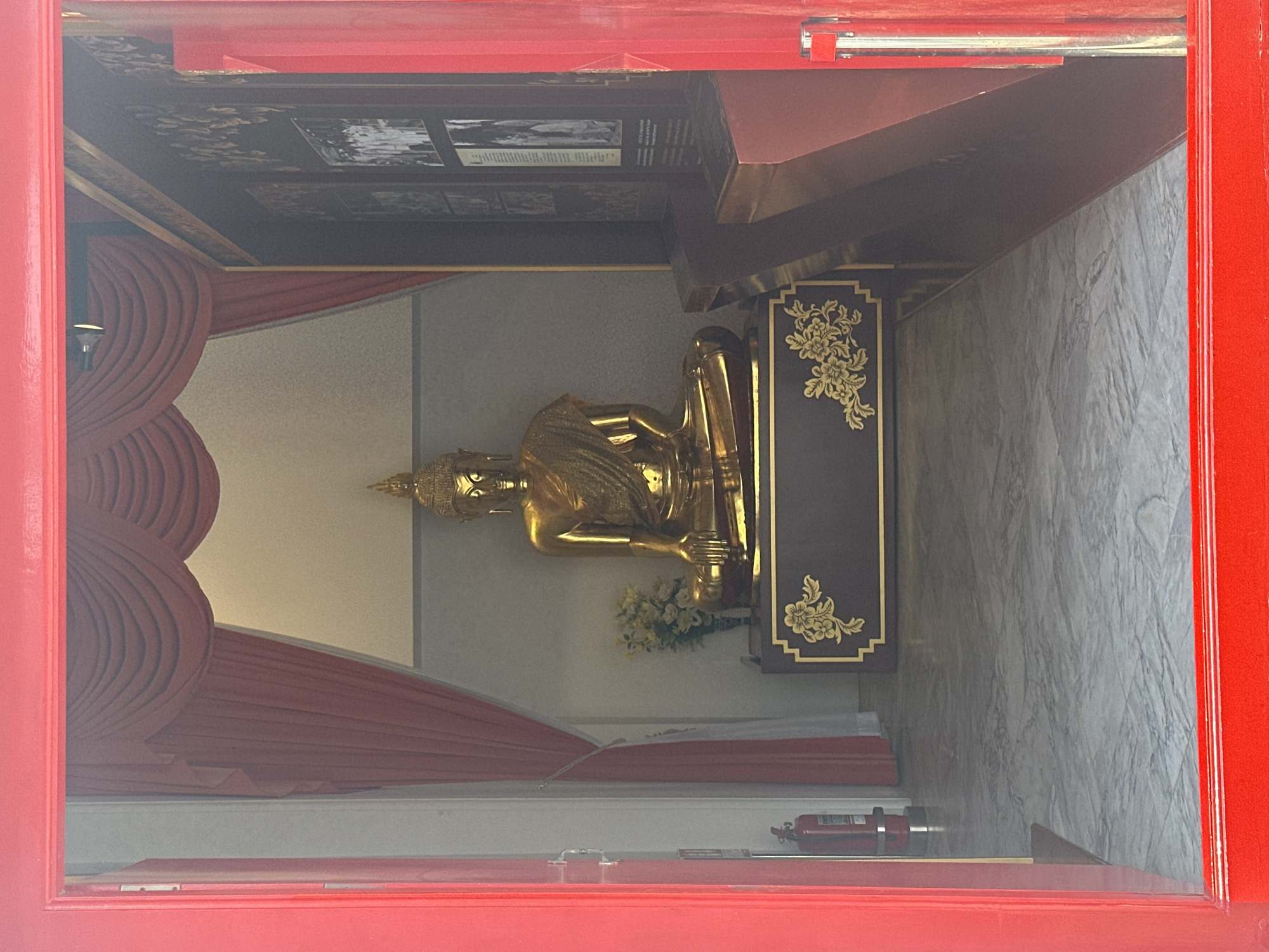 A golden Buddha statue is the focal point, viewed through a red-framed window. The statue is positioned on a dark brown base adorned with gold floral designs. A small bouquet of flowers sits at the statue's base. Red curtains are draped behind the statue, contrasting with the light gray walls. The setting appears to be inside a temple, potentially in Bangkok, Thailand.