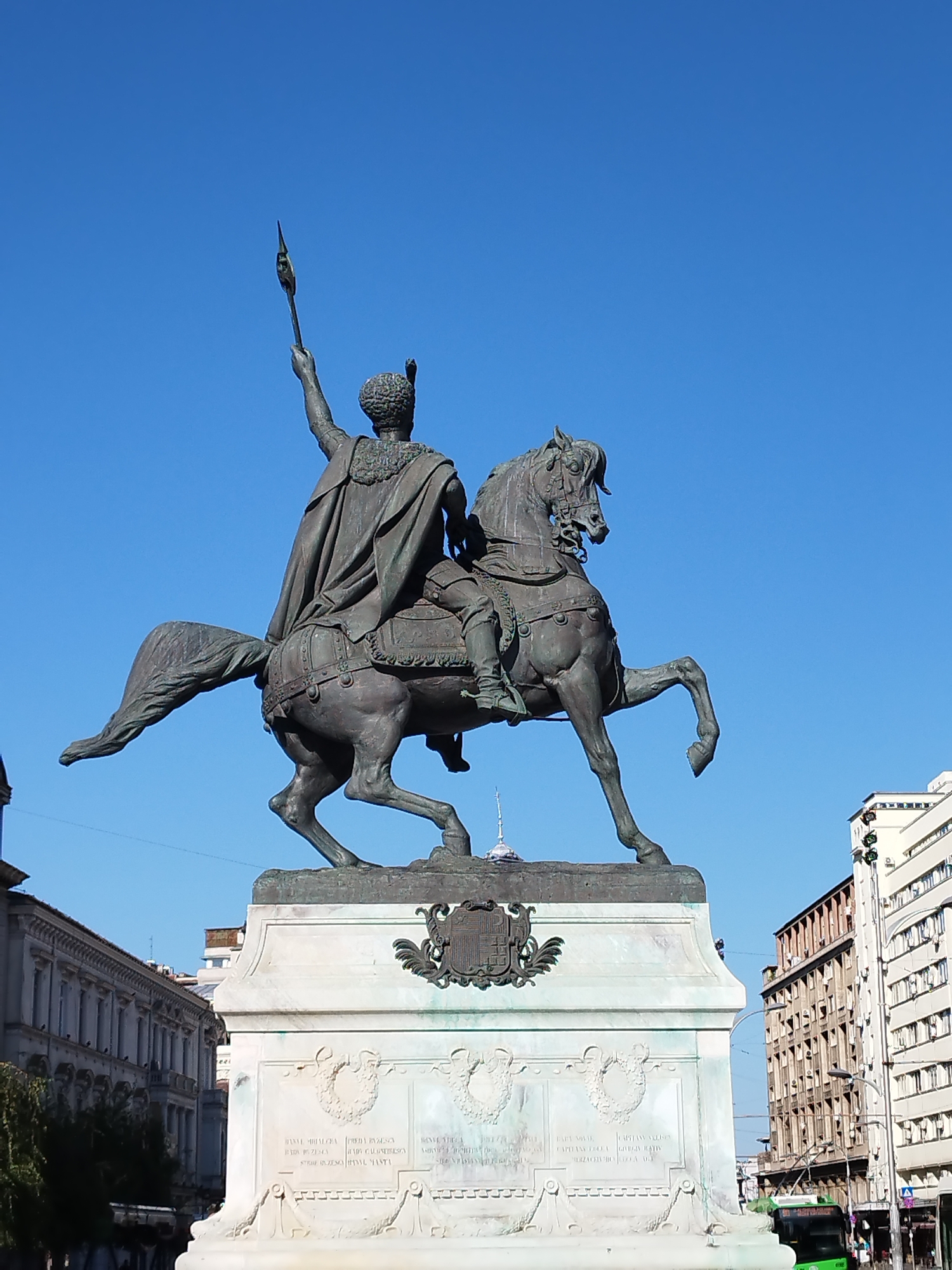 The image depicts a bronze equestrian statue of King Michael I of Romania, located in Bucharest, Romania. The statue features the king mounted on a horse, with his right arm raised, holding a spear. He is dressed in a cape and armor. The statue stands on a large white marble pedestal adorned with relief carvings and inscriptions. In the background, several buildings are visible, characteristic of an urban environment, with a clear blue sky above. A green bus is visible on the right side of the image, suggesting ongoing city traffic. The statue is a prominent landmark in Bucharest, situated in a public square.