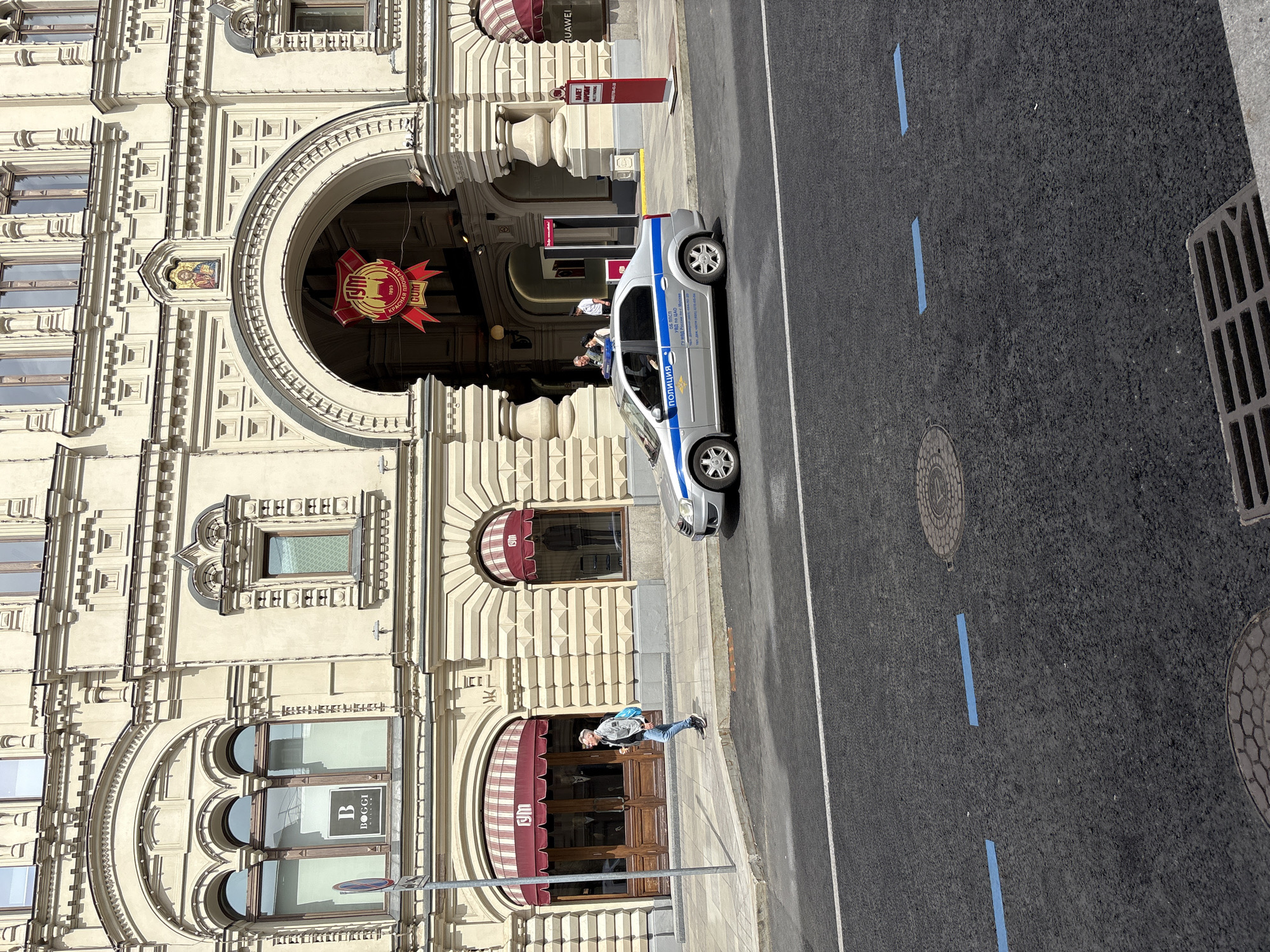 The image captures a scene in an urban location, featuring a cream-colored building with ornate architectural details, likely a commercial or historical landmark. Red-striped awnings cover the windows and doorways of the building. A silver police car is parked on the asphalt road alongside the building. The road has white lines and blue markers indicating traffic lanes.
A person is visible walking on the sidewalk in front of the building. A red and yellow sign is visible above the door to the building. A Boggi Milano logo is visible in the lower left of the image. A red and yellow sign is visible near the top of the building under the archway. A road drainage grate can be seen in the lower right corner of the image.