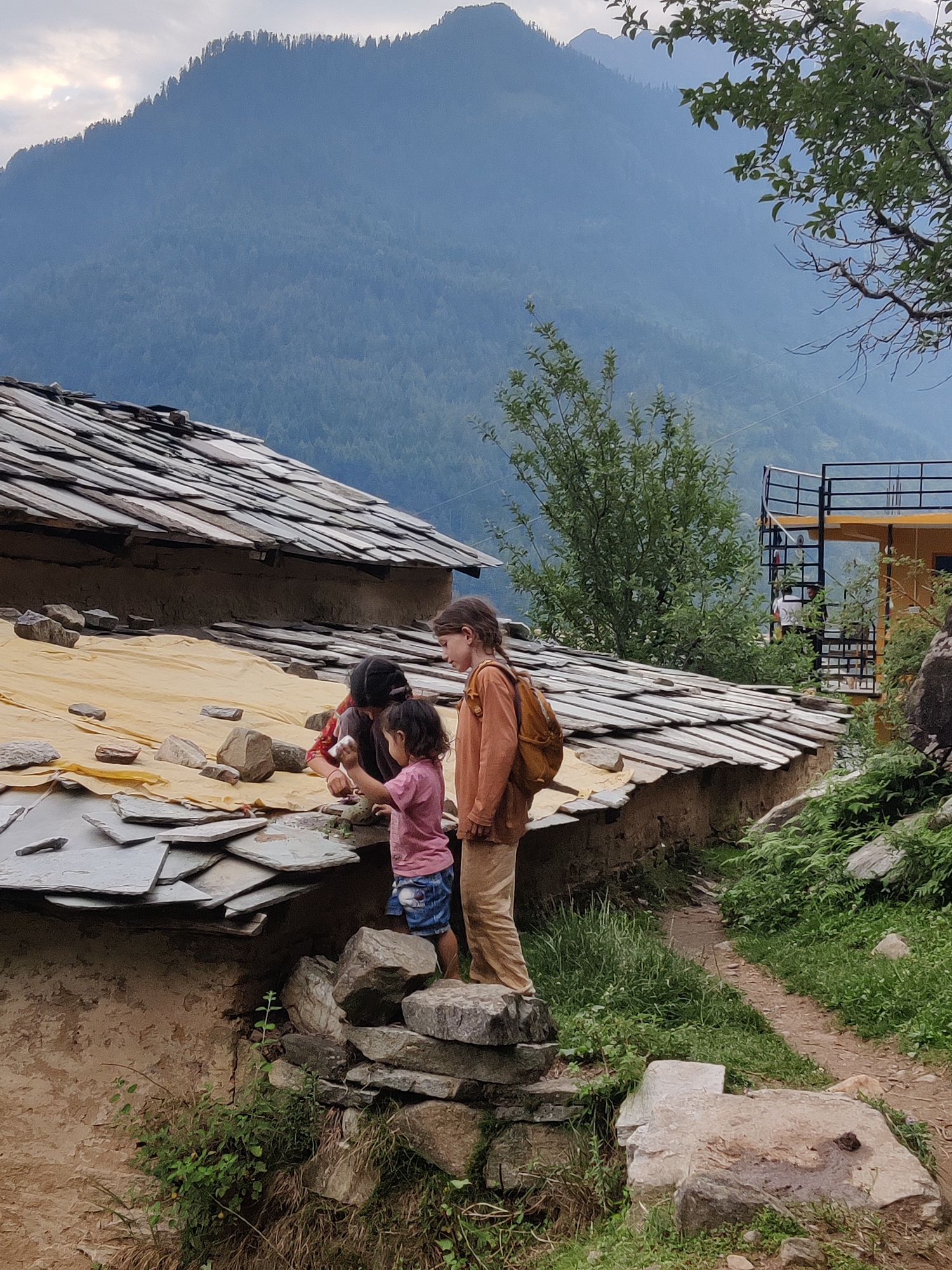 In the village of Vashisht, India, three children are gathered around a partially covered roof of a building constructed with natural materials. The roof is made of slate tiles, with a yellow tarp visible underneath in some sections. The children are actively engaged with the roof; one older child, wearing an orange shirt and a brown backpack, stands to the right, observing. Two younger children are crouched down, interacting with the slate tiles and the tarp. One child, wearing a pink shirt and blue shorts, is holding a white object, possibly a tool or a piece of material, and seems to be placing something onto the roof. The other child, wearing a red and black patterned top, is also leaning over the roof, looking down. The background features a lush green hillside with trees, and a large, hazy mountain dominates the upper portion of the image. The overall scene depicts a moment of rural life, with children contributing to or interacting with the infrastructure of their home.