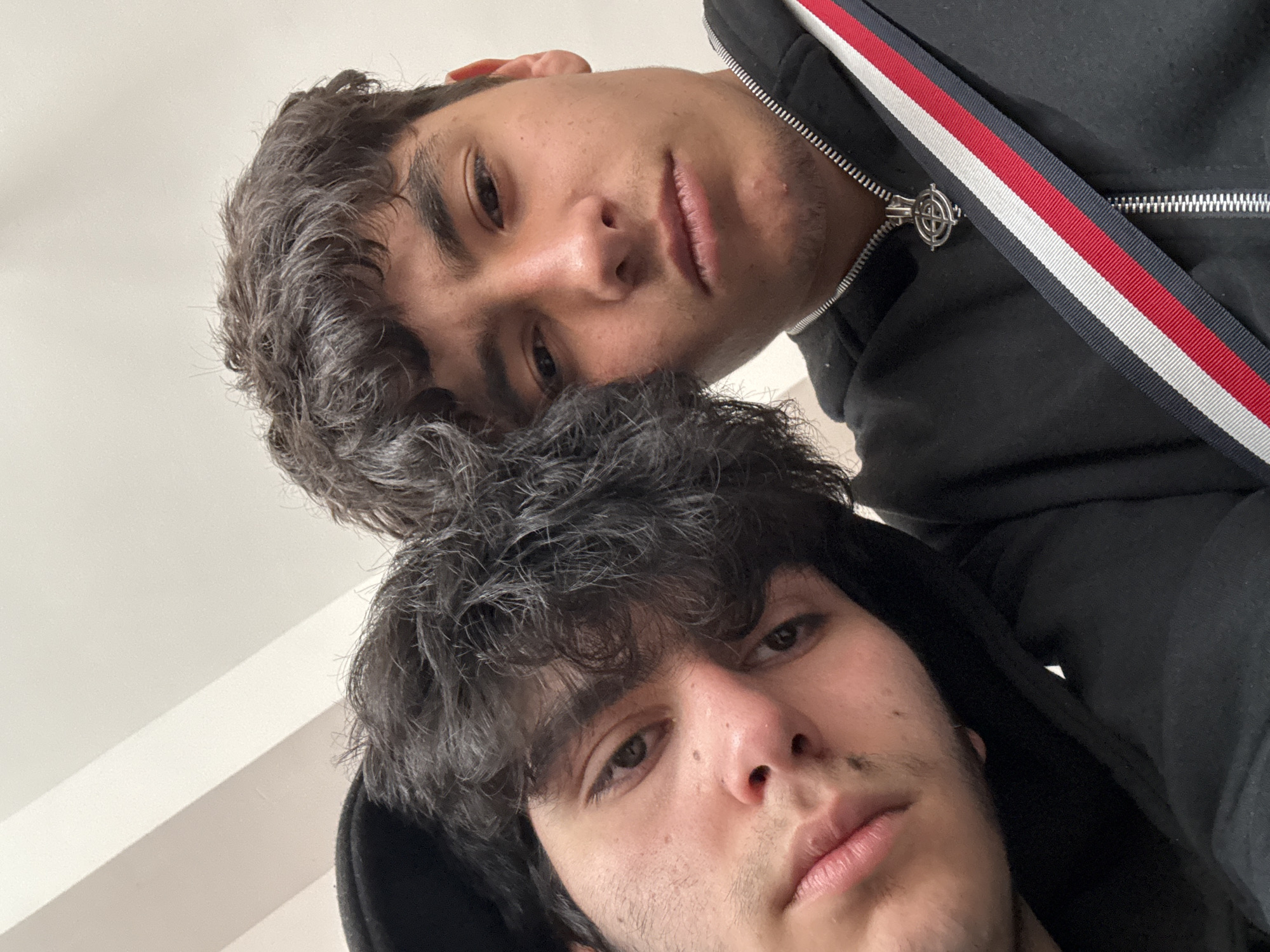 Two young men pose closely together for a selfie. The upper individual wears a black hoodie jacket with a red, white, and blue striped detail along the zipper line, while the other is in a black hoodie. The background appears to be a plain, white ceiling with a slight angular architectural element. The location of the photo cannot be determined based on the provided image alone.