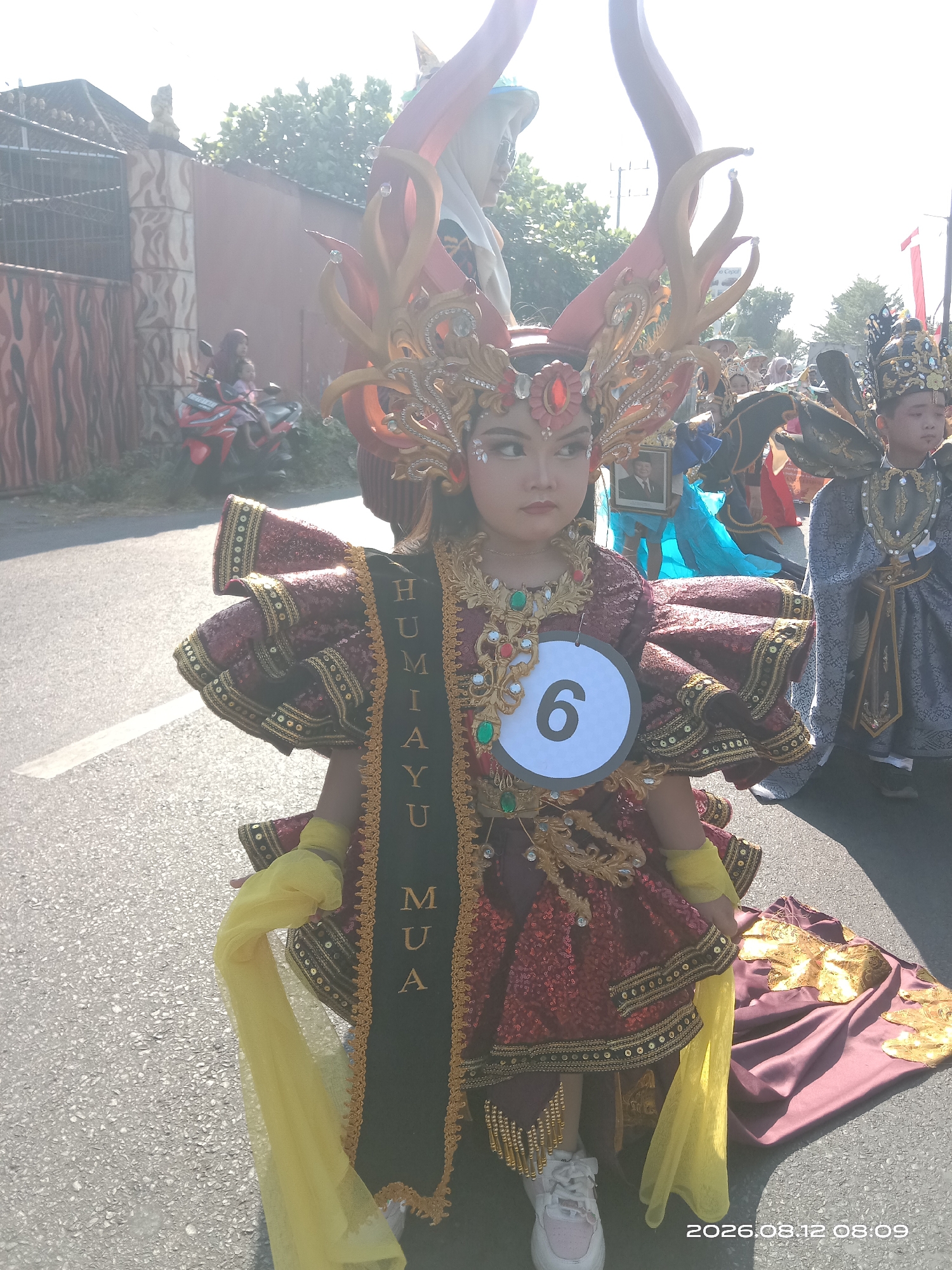 In Gudo, Indonesia, a young girl dressed in a vibrant, traditional costume participates in a parade. The costume features a maroon sequined dress with ruffled sleeves, adorned with intricate gold embellishments and a sash that reads "HUMIAYU." She wears a large, ornate headpiece resembling antlers and a crown, decorated with red and gold accents and crystals. A white circle with the number "6" is pinned to her chest. Yellow fabric drapes from her sleeves and a matching train lies on the ground. She is surrounded by other participants in similarly elaborate attire. In the background, a stone wall with an arched gateway and some trees are visible, and a motorcycle with two passengers is parked nearby. The ground is paved with asphalt. The timestamp on the image reads "2026.08.12 08:09".