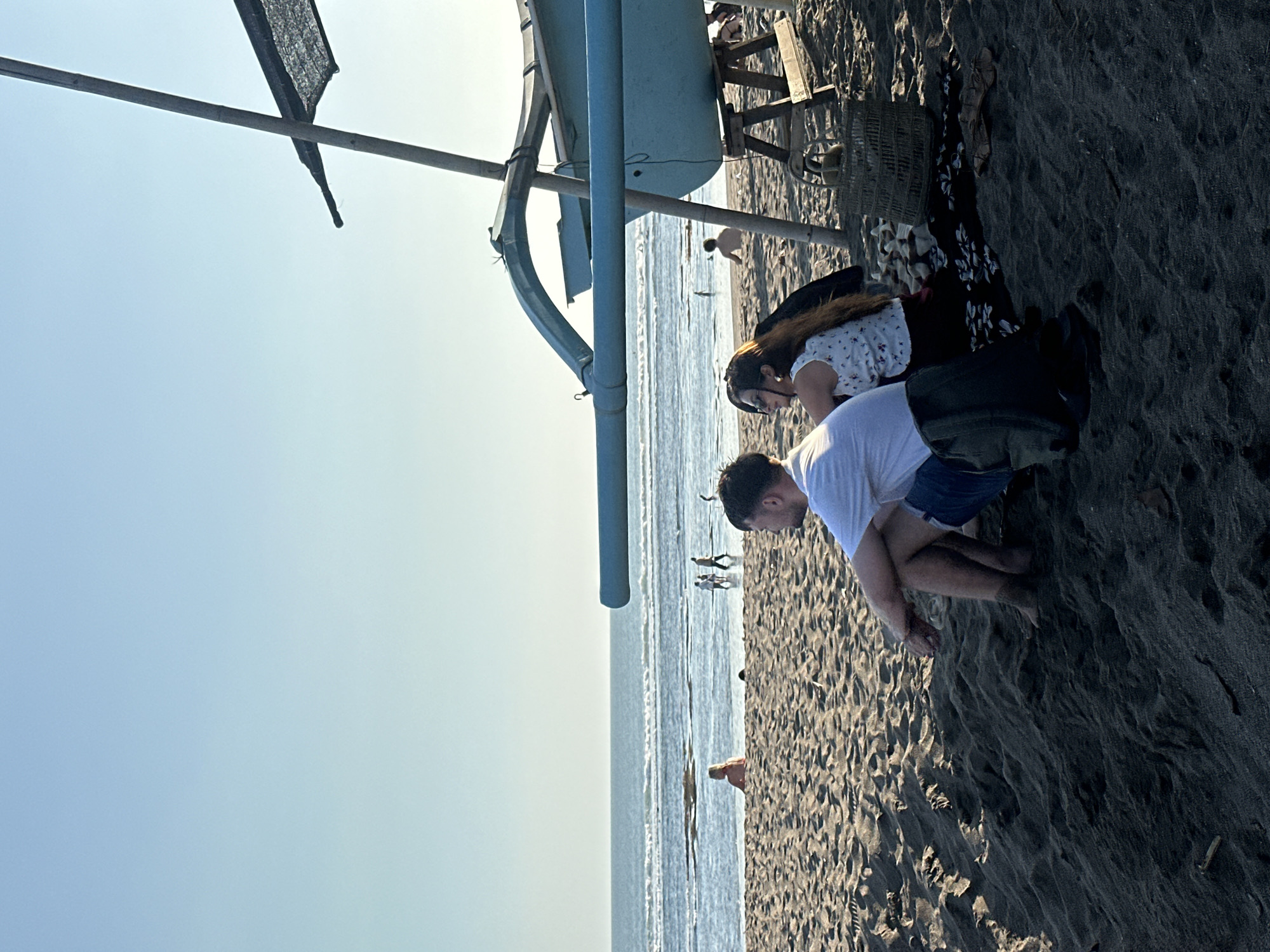 On a beach in North Kuta, Indonesia, a man and a woman are seated on dark sand, facing the ocean. The man, wearing a white t-shirt and blue shorts, is in the foreground, his legs crossed. The woman, with long brown hair, is to his right, wearing a white and red floral top and a dark skirt. They are seated near a light blue outrigger canoe propped up on wooden stands. The canoe has a bamboo pole and a dark net stretched over it. In the background, the ocean has small waves, and several people are visible in the water and walking along the shoreline. The sky is clear and blue.