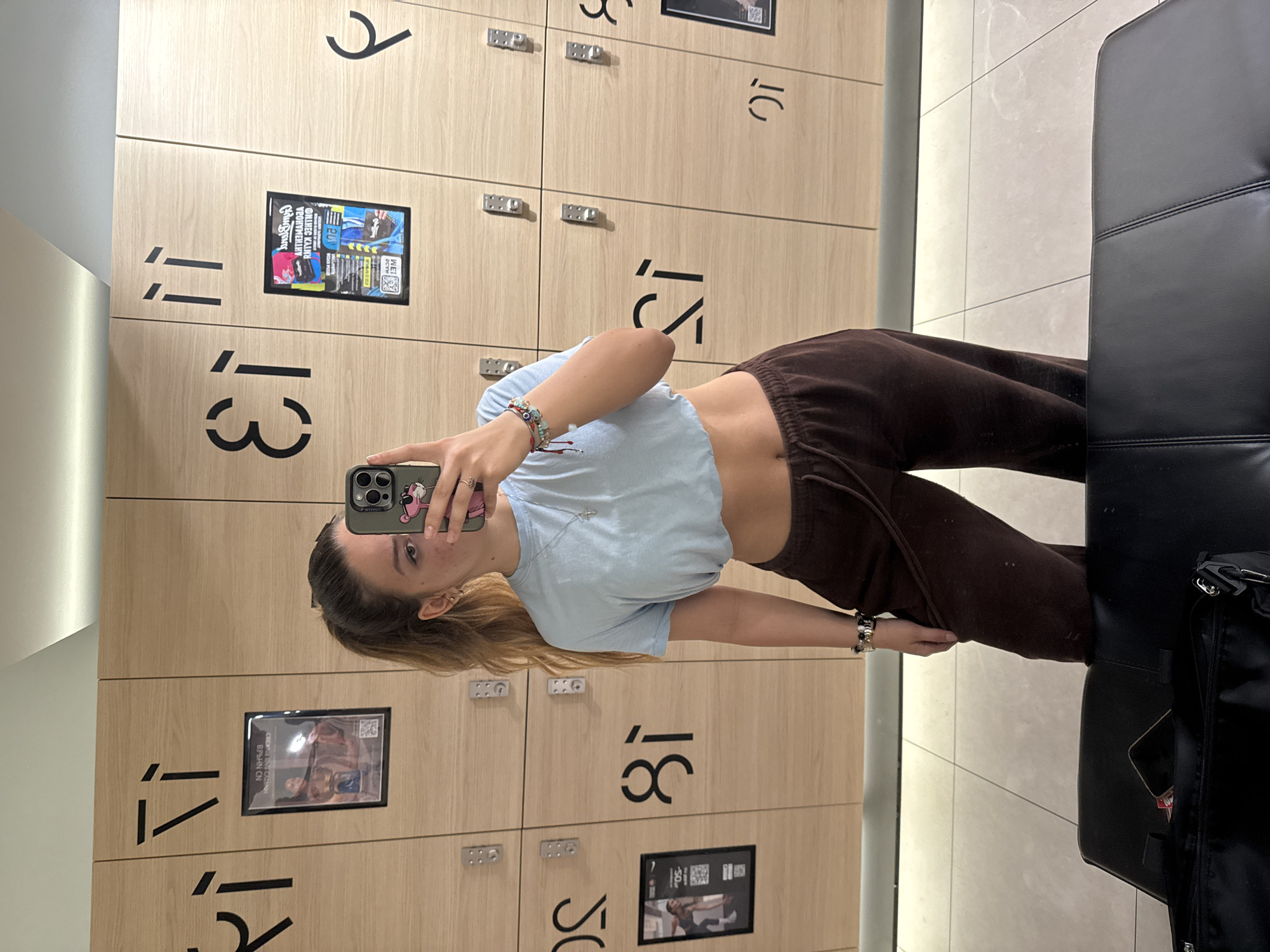 The image depicts a mirror selfie of a young woman taken in the locker room of a Pulsar Fitness gym located in Sofia, Bulgaria. The woman has light brown hair tied in a high ponytail and is wearing a light blue cropped t-shirt, dark brown sweatpants, and multiple bracelets on her left wrist. She holds a dark-colored smartphone with a pink panther sticker, partially obscuring her face, and has her tongue slightly extended. The background consists of light wood-paneled lockers, each equipped with a metal key lock and stylized black numbers or letters. Visible unmirrored locker identifiers include "71," "31," "11," "05," "18," "51," and "01." Several lockers display framed posters containing fitness-related imagery and mirrored Bulgarian text, notably "АЕРОБНИ И СИЛОВИ ТРЕНИРОВКИ" (Aerobic and Strength Workouts), confirming the gym's national context. The floor is composed of light-colored tiles. In the foreground, a black faux leather bench is visible, supporting a black duffel bag and another smartphone. The scene is evenly lit by overhead artificial lighting.