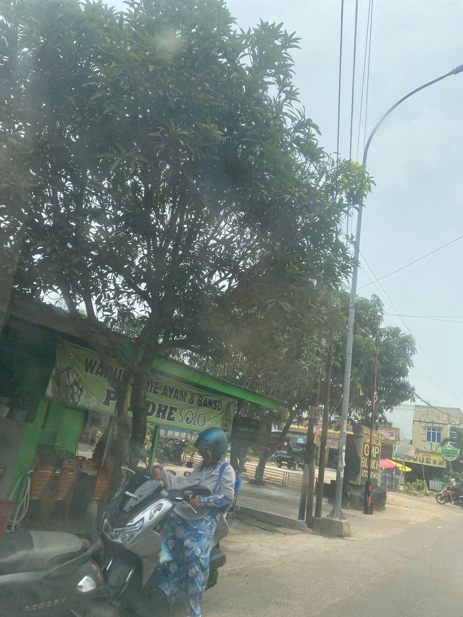 An outdoor, daytime scene features a woman wearing a blue helmet and a patterned blue and white outfit standing next to a silver scooter on a paved road. She is holding the scooter's handlebars. Behind her, a green building with a banner that reads "WARUNG PIE AYAM & BAKSO DHE SOLO" is visible. Several chairs are stacked on the side of the building. Trees with dense green foliage line the area. Overhead, power lines and a street light are present. In the background, other buildings and vehicles can be seen, including a truck with "MITSUBISHI" on its side and a person on a motorcycle. The sky is overcast. The text on the banner suggests the location is likely in or around Solo, Indonesia, rather than Batam.
