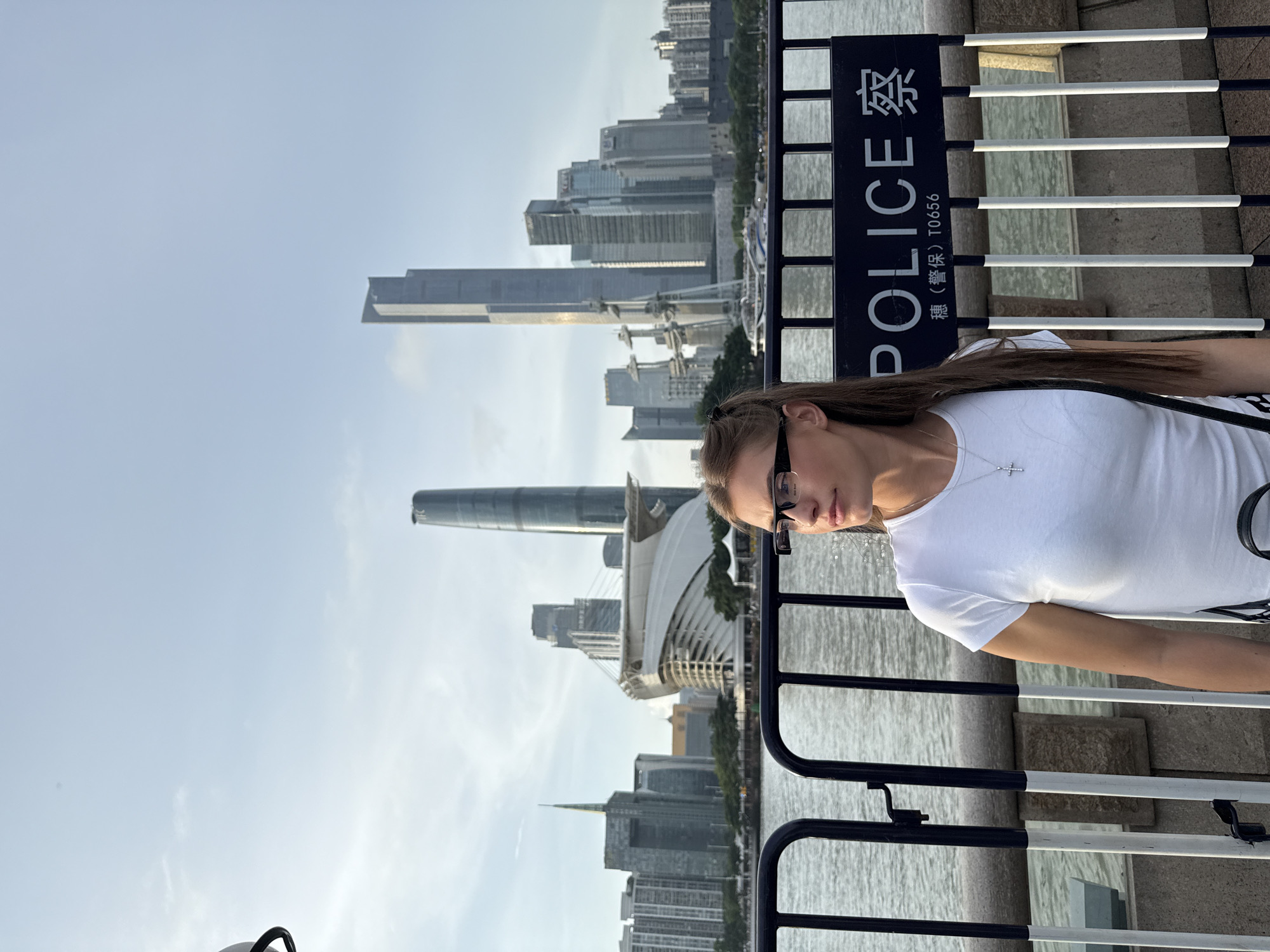 A woman with long brown hair and glasses stands in front of a modern cityscape. She is wearing a white t-shirt and a silver cross necklace. She is looking to her left, with a neutral expression. In the background, a body of water is visible, with a dense collection of tall buildings and skyscrapers lining the opposite bank. A prominent sign is to her right, which reads "POLICE" in English and also contains Chinese characters and the text "穗(警保) T0656". The scene appears to be set during the daytime, under a clear sky with some scattered clouds. The overall atmosphere is urban and contemporary, with a focus on the architectural landscape.
