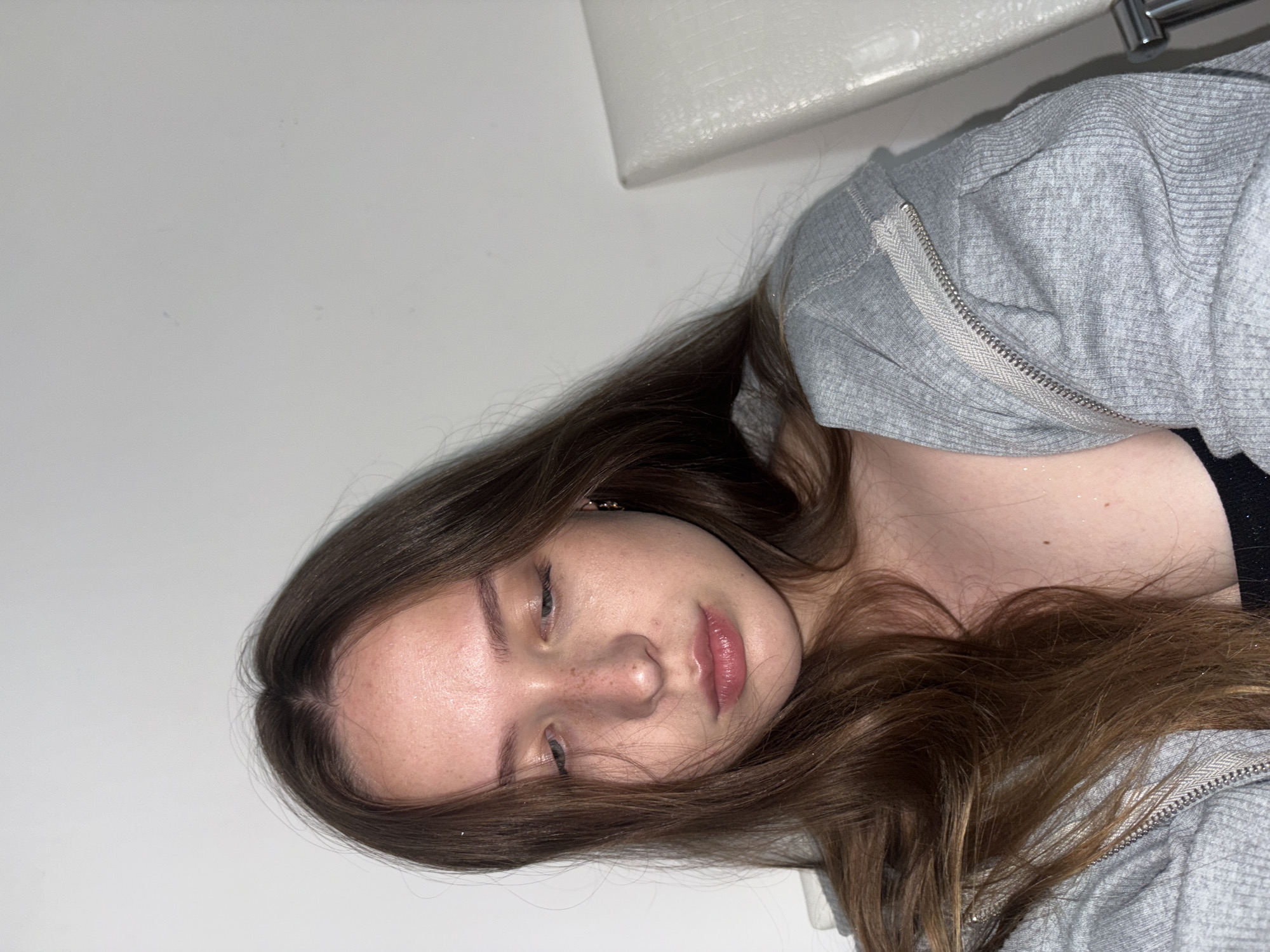 A young woman with long, brown hair is shown from the chest up, looking downwards and to her right. She has fair skin with freckles on her nose and cheeks, and she is wearing a grey zippered hoodie over a black top. The background is a plain white wall, with a portion of a white textured chair visible on the right side of the frame. The image appears to be taken indoors, with a slightly overhead perspective. There is no information in the image to indicate the location in the city of Chaikovsky, Russia.
