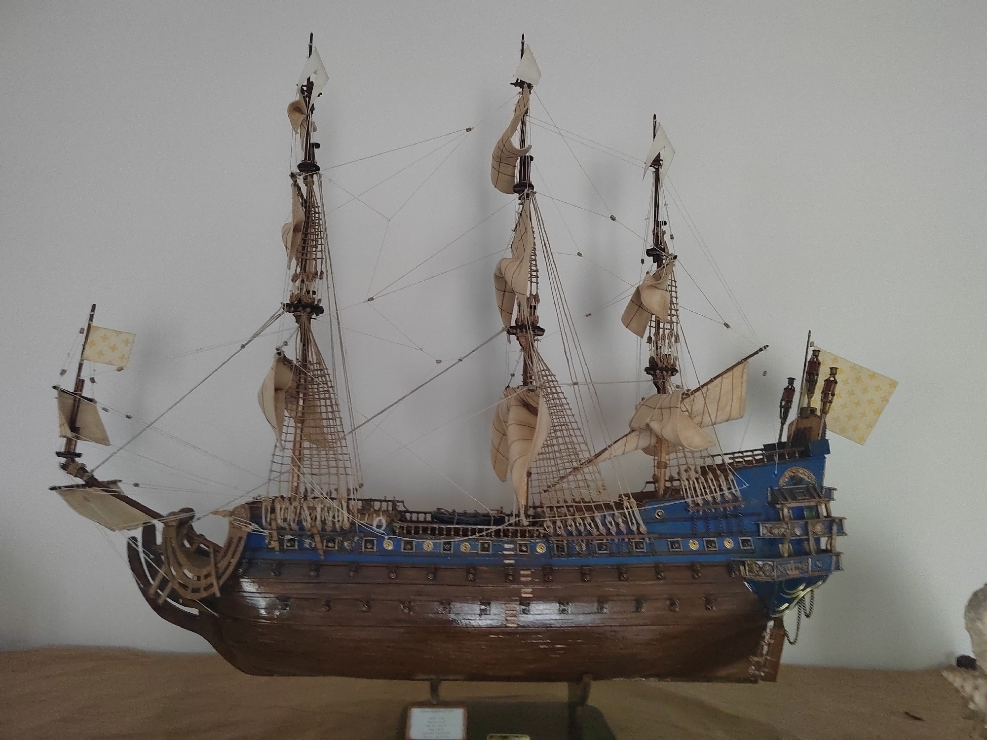 This is a detailed, front-facing photograph of a wooden model ship displayed against a plain, off-white background. The ship is intricately constructed, featuring multiple masts, sails, rigging, and decks. The hull is primarily a dark brown wood with a glossy finish, while the upper portion of the hull and stern are painted a vibrant blue. The ship is equipped with several flags; a prominent one on the starboard stern is yellow with a repeating pattern of gold fleur-de-lis. Another smaller flag, also yellow with a fleur-de-lis pattern, is visible on the port bow. The ship is mounted on a stand with dark supports. In the foreground, a light brown, textured surface, likely a table or shelf, is visible. There is a small, rectangular white label on the stand in front of the ship, which is illegible. The lighting appears to be from above, casting slight shadows. There are no people visible in the image. The scene is static and does not depict any action. The location of this model ship within Porto Recanati, Italy, is not discernible from the visual information provided in the photograph.