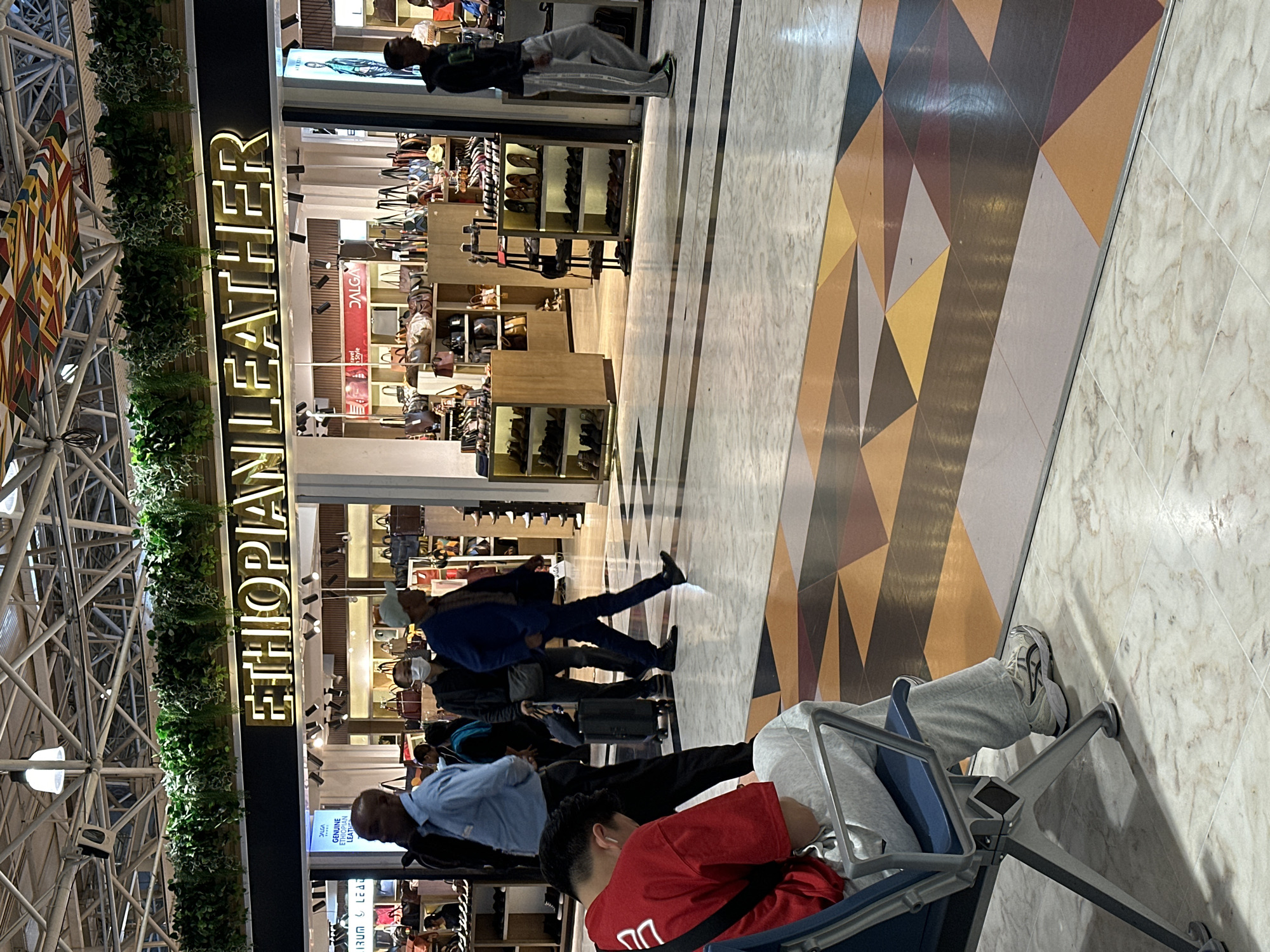 The image depicts the interior of a commercial establishment, likely a store, with the signage "ETHIOPIAN LEATHER" prominently displayed in illuminated golden lettering against a black background. The store appears to specialize in leather goods, with shelves and displays visible behind the signage showcasing numerous bags and possibly shoes. The environment suggests an indoor shopping area, possibly within an airport or a mall, given the polished floor with geometric patterned tiles.
In the foreground, a young person wearing a red shirt and gray sweatpants is seated on a blue and metal chair, facing away from the camera and appearing to be looking down, possibly at a phone or the floor. Their feet, clad in white athletic shoes with black accents, rest on the tiled floor.
Several other individuals are visible in the mid-ground. Two men in professional attire, one in a light blue shirt and the other in a blue suit, are walking. Another man, wearing a face mask, is also walking, pulling a small rolling suitcase. Further in the background, more shoppers and staff are present, with various merchandise displayed. The overall lighting is bright, suggesting artificial illumination. The scene conveys a sense of movement and commerce within an indoor public space. The location is the Ethiopian Leather shop at Bole International Airport in Addis Ababa, Ethiopia.