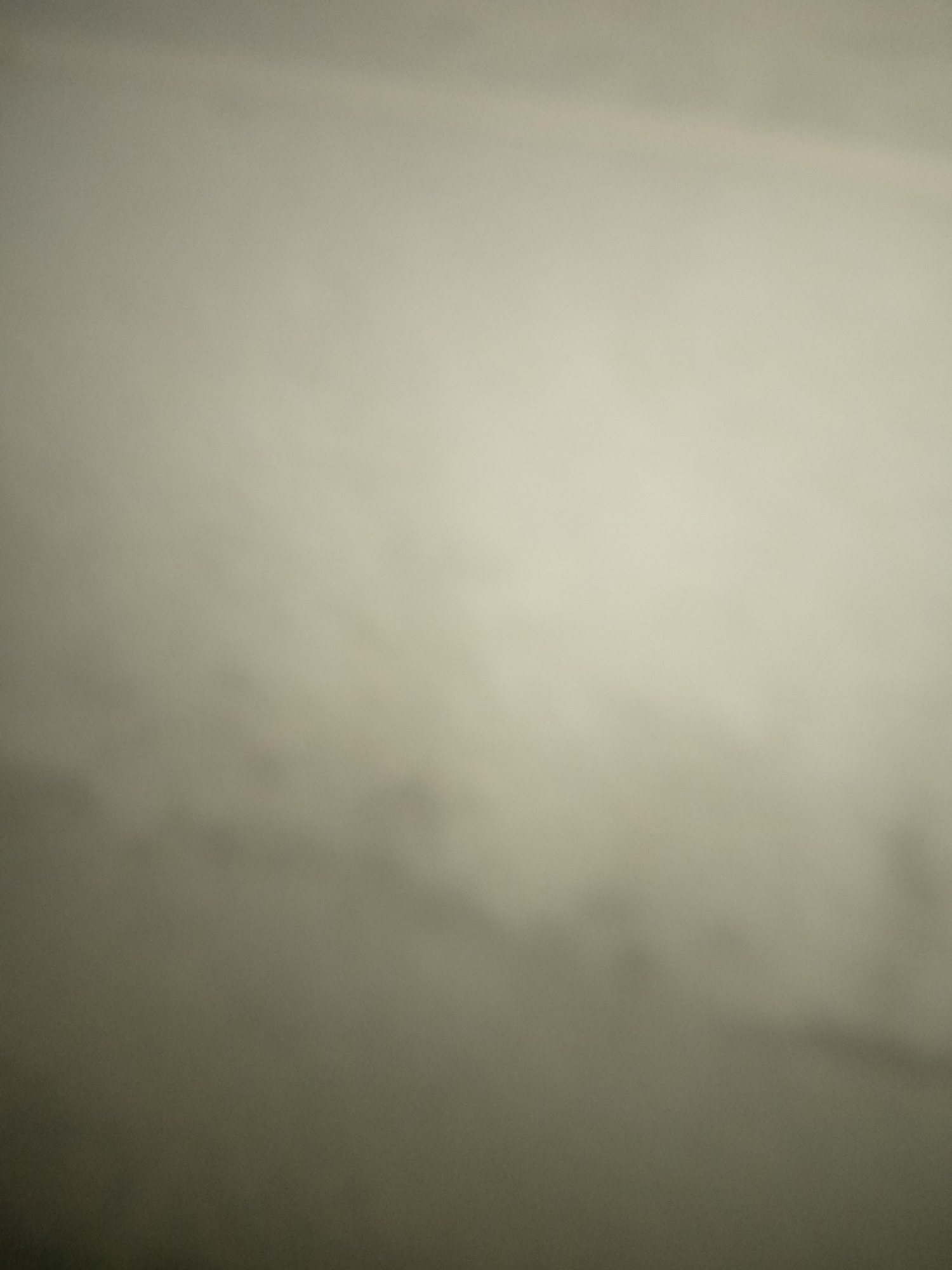 The image is a blurred, abstract photograph of a textured surface, possibly a wall or fabric. The colors are muted, ranging from a light beige to a darker, shadowy gray, with a gradient effect that suggests a light source is present. There are subtle, darker splotches or patterns visible in the lower half of the image, creating a sense of depth and texture. The overall impression is one of softness and diffused light, with no discernible objects or figures. The location is not specified in the image.