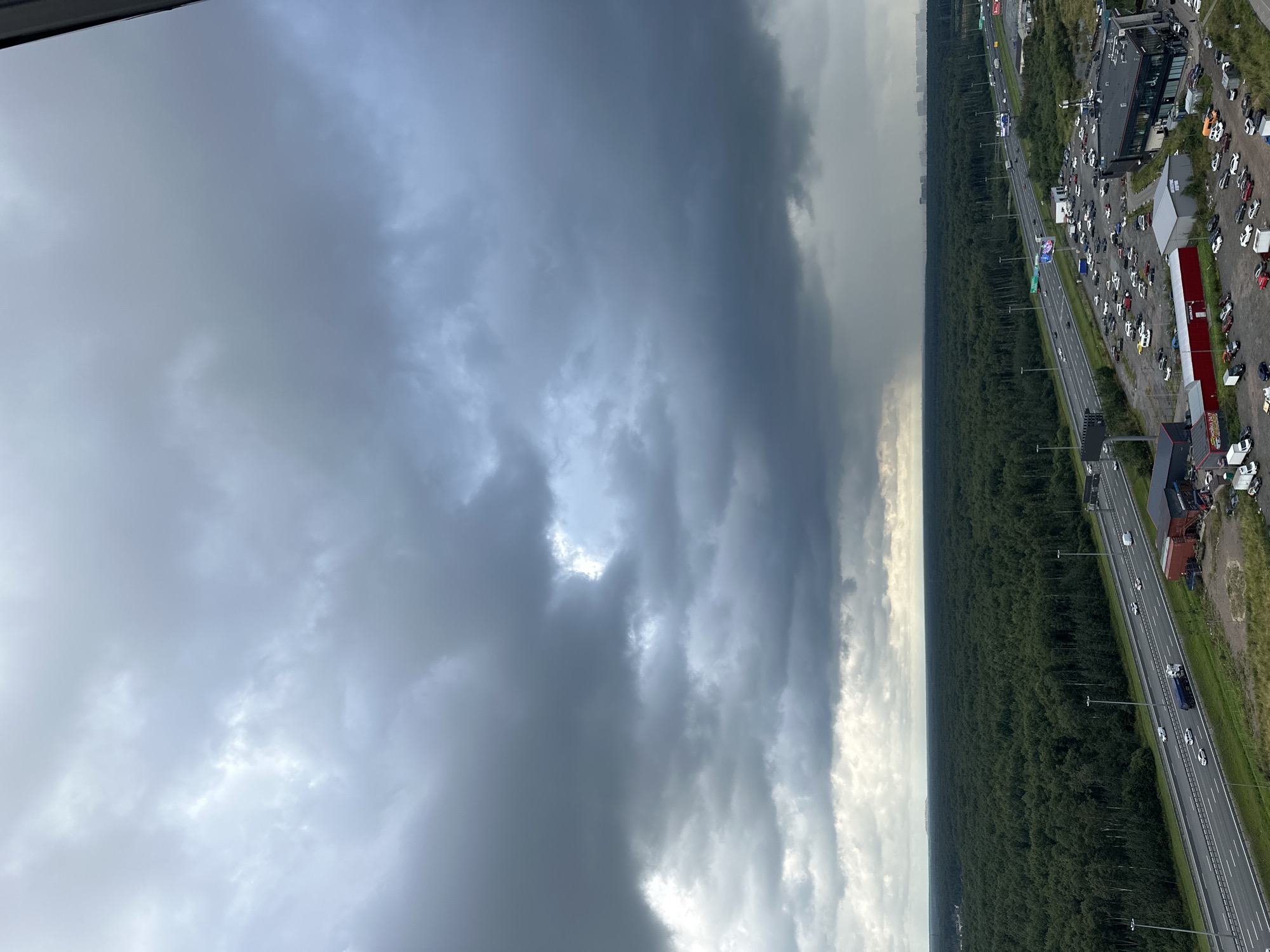 The image depicts an aerial view of a highway passing through a dense forest near Otradnoye, Russia. Dark, stormy clouds dominate the sky, with a distinct area of rain visible in the distance. The highway is lined with streetlights and is populated by several vehicles, including cars and trucks. Adjacent to the highway, there is a large parking lot filled with numerous cars, and several commercial buildings, one of which is a modern structure with a dark facade. The forest extends to the horizon, and in the far distance, a city skyline is faintly visible through the atmospheric haze.