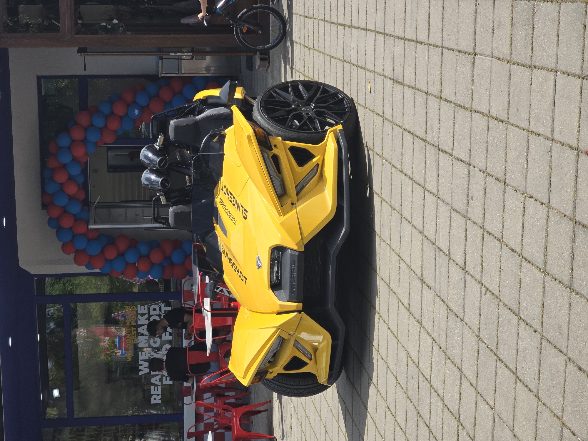 The image depicts a yellow Polaris Slingshot parked on a paved area, likely a sidewalk or parking lot. The Slingshot is facing away from the camera at a slightly angled position. Behind the vehicle, the facade of a building is visible, decorated with an arch of red and blue balloons. The building has glass windows and a sign is visible. Red chairs and tables are set up near the entrance of the building. A bicycle is visible in the background. Two people are seen inside the building. The environment is sunny and outdoors. The image does not specify the city.