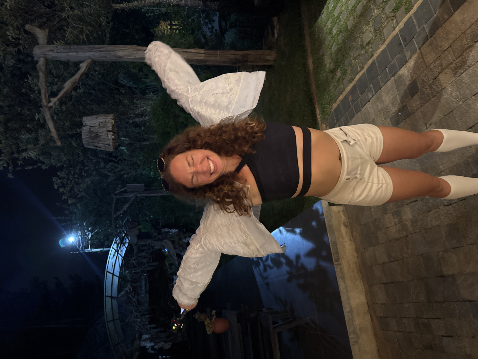 A woman with curly brown hair and a closed-mouth smile is standing outdoors at night with her arms outstretched, her white ruffled sleeves resembling wings. She is wearing a black crop top with a criss-cross neckline, white shorts, and white knee-high socks. She has sunglasses perched on her head. The background features dark trees, a pathway made of dark stones, and some outdoor furniture. The scene appears to be in a semi-rural or garden setting, possibly a restaurant or resort, in the city of Moc Chau, Vietnam. A bright light is visible in the upper left portion of the image.