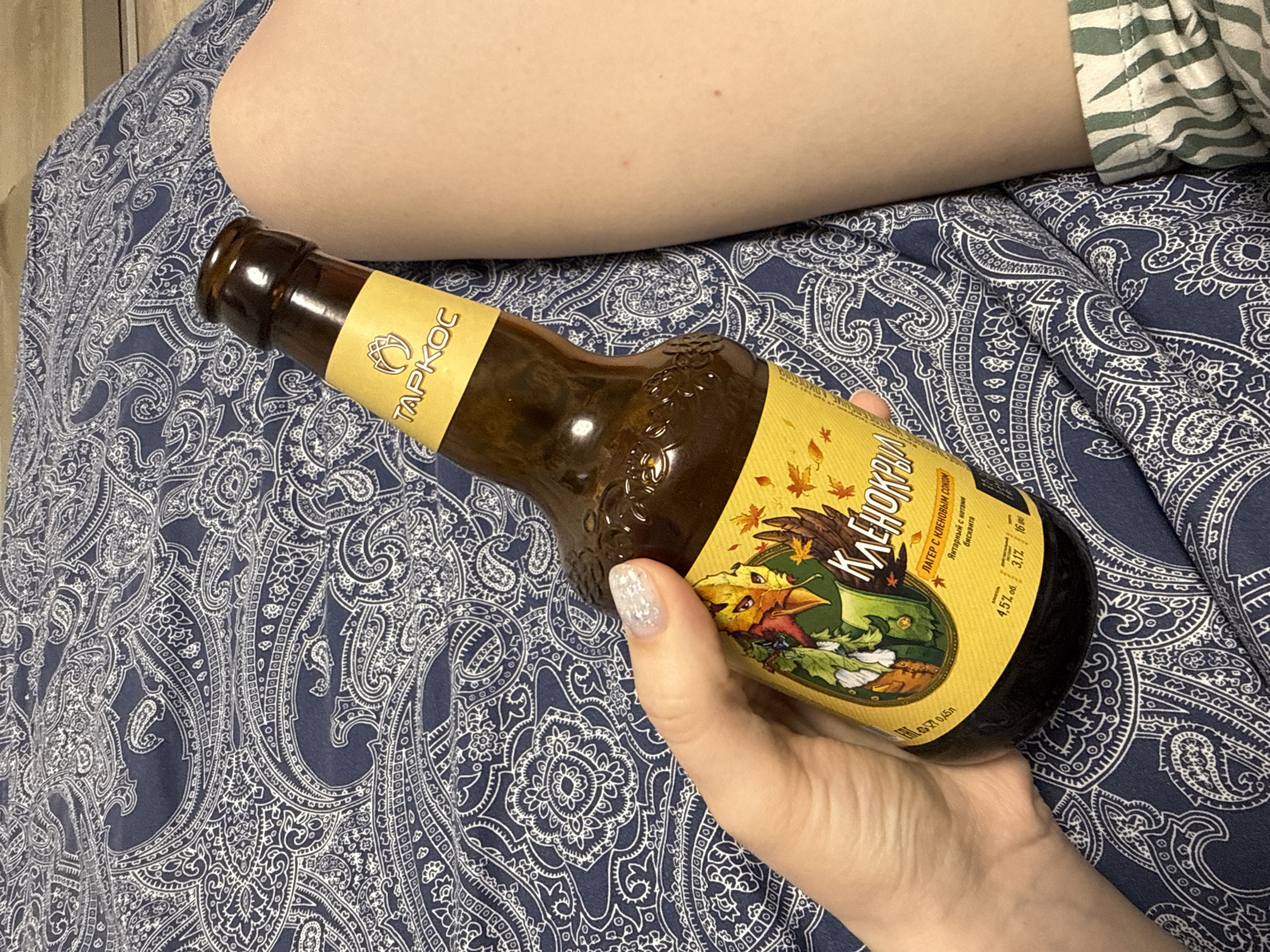 A hand is holding a brown glass bottle of "Klenokryly" beer, a lager with maple sap, produced by TAPKOC, which is written in Russian. The beer is described as amber with notes of biscuit, with 4.5% alcohol and 3.1% density. The background is a patterned blue and white fabric, possibly a bedspread. A human thigh is partially visible at the top of the image. The context indicates it is in Russia, possibly Moscow, due to the Russian Cyrillic writing on the bottle.