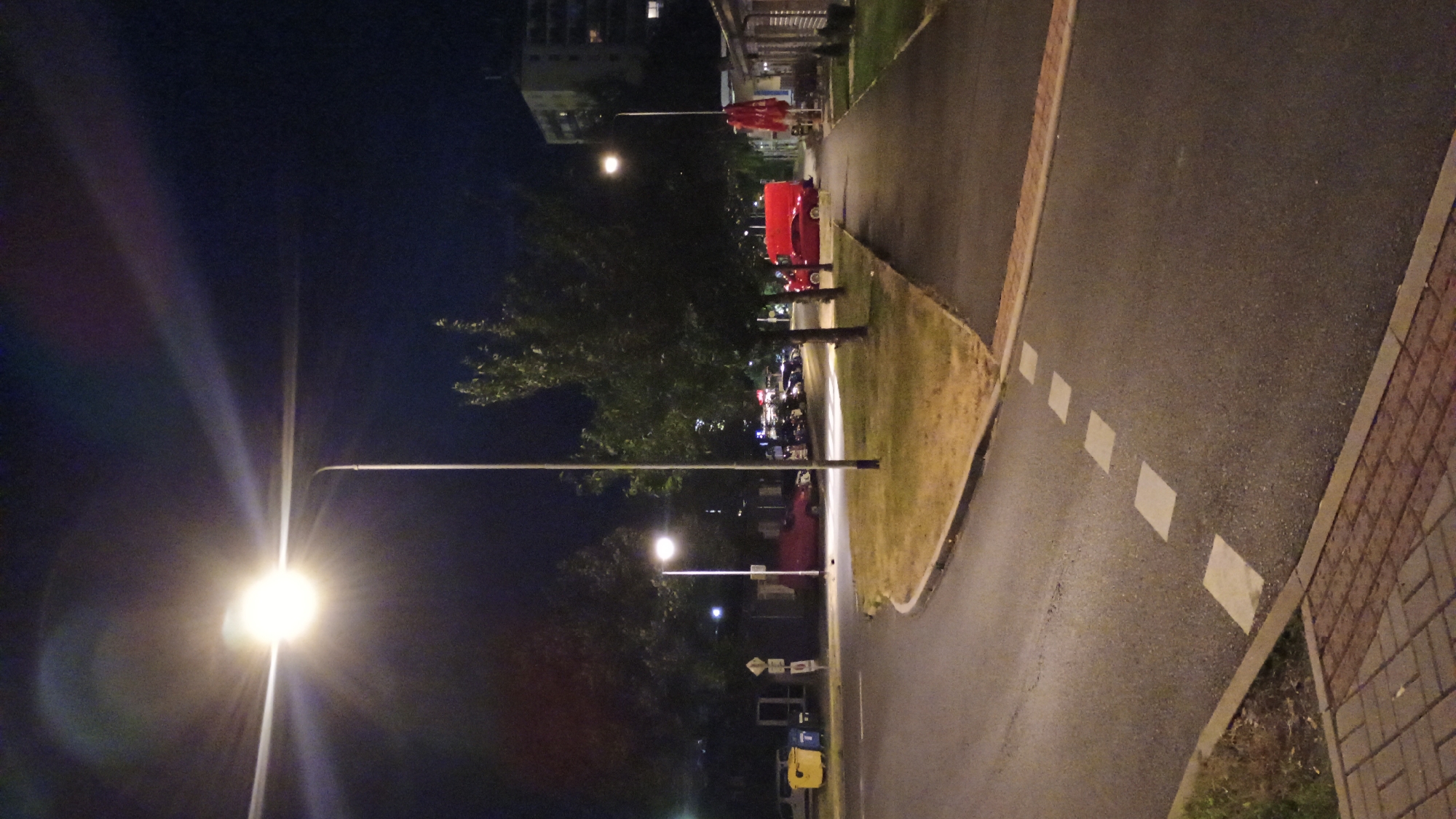 The image depicts a street scene at night in Sokolov, Czechia. A bright streetlamp illuminates the foreground, casting a strong glare. The street itself is paved, with white square markings indicating a pedestrian crossing or lane division. To the left, a curved road leads into the distance, bordered by dry grass and trees. Several parked cars are visible, including a red van and a red car. A building with lights on is also present in the background. A sidewalk runs along the right side of the street, adjacent to a row of low buildings, one of which has a red umbrella outside. The sky is dark, with the bright streetlamp being the dominant light source. There are no people visible in the image.