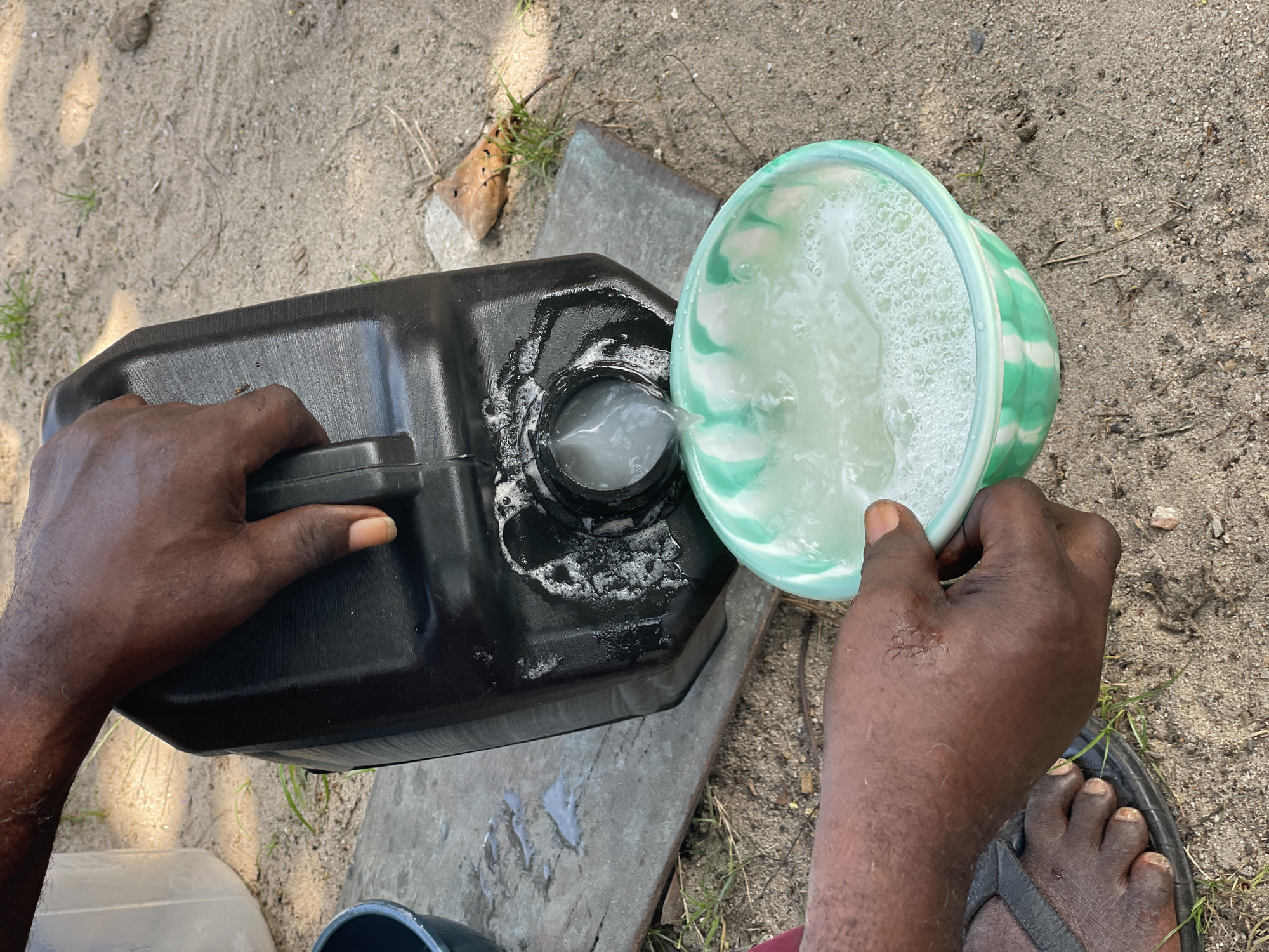 An overhead shot captures a person pouring a milky white, frothy liquid from a black, rectangular jerrycan or large plastic container into a green and white patterned plastic bowl. The person's dark-skinned hands are prominent; the left hand grips the handle of the black container, tilting it, while the right hand steadies the green bowl.
The liquid, which appears opaque and foamy, flows from the opening of the jerrycan directly into the bowl, which already contains a similar-looking liquid with a bubbly surface. The ground beneath the activity is sandy, light brown earth, sparsely covered with small patches of green grass and a few dry leaves or debris. A grey concrete slab is visible beneath the containers. Part of a bare right foot, wearing a black flip-flop, is visible in the lower right corner of the frame.
The scene is brightly lit, indicating daytime. There is no discernible text on any of the objects. The image depicts a practical task, likely an outdoor chore in a location like Badagry, Nigeria, where such containers are commonly used for transporting and dispensing liquids.