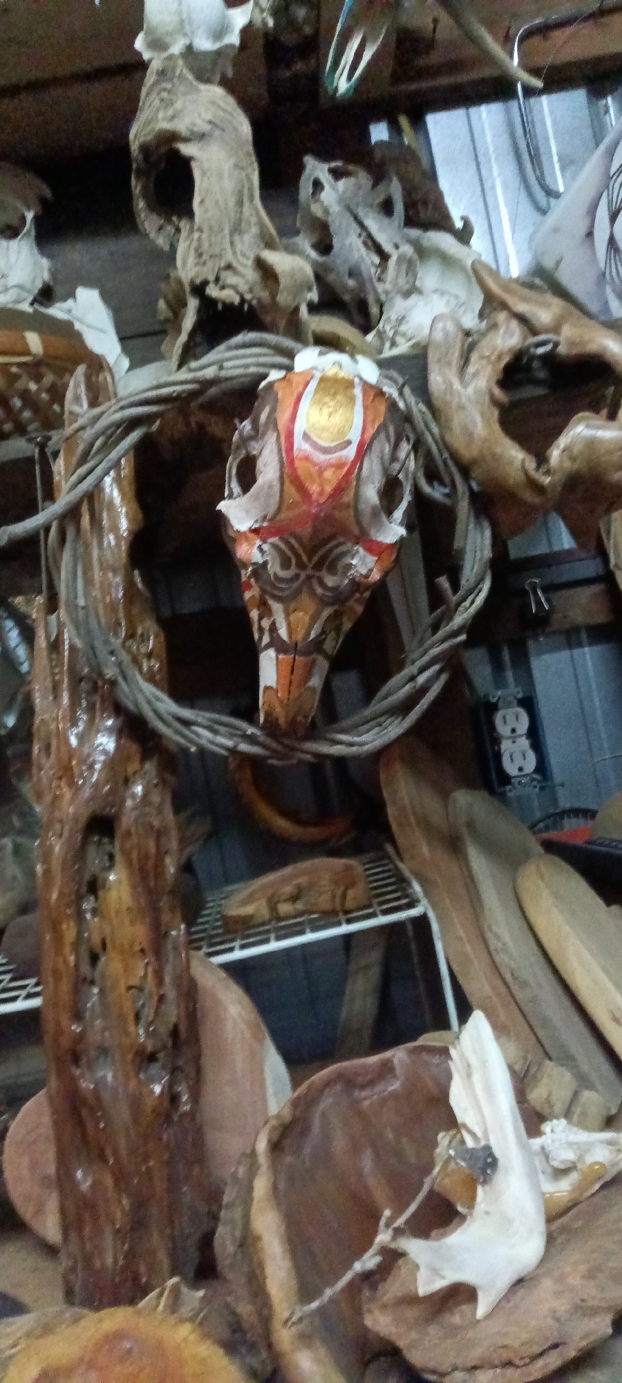 The image depicts a collection of decorative items, primarily consisting of wooden objects and animal skulls, possibly in a shop or workshop. A painted animal skull, adorned with orange, white, and gold designs, is centrally positioned and encircled by a woven wreath of twigs. Other wooden pieces, including what appear to be driftwood and carved elements, surround the skull. Electrical outlets are visible on the wall in the background. The location could be in Tahlequah, United States.